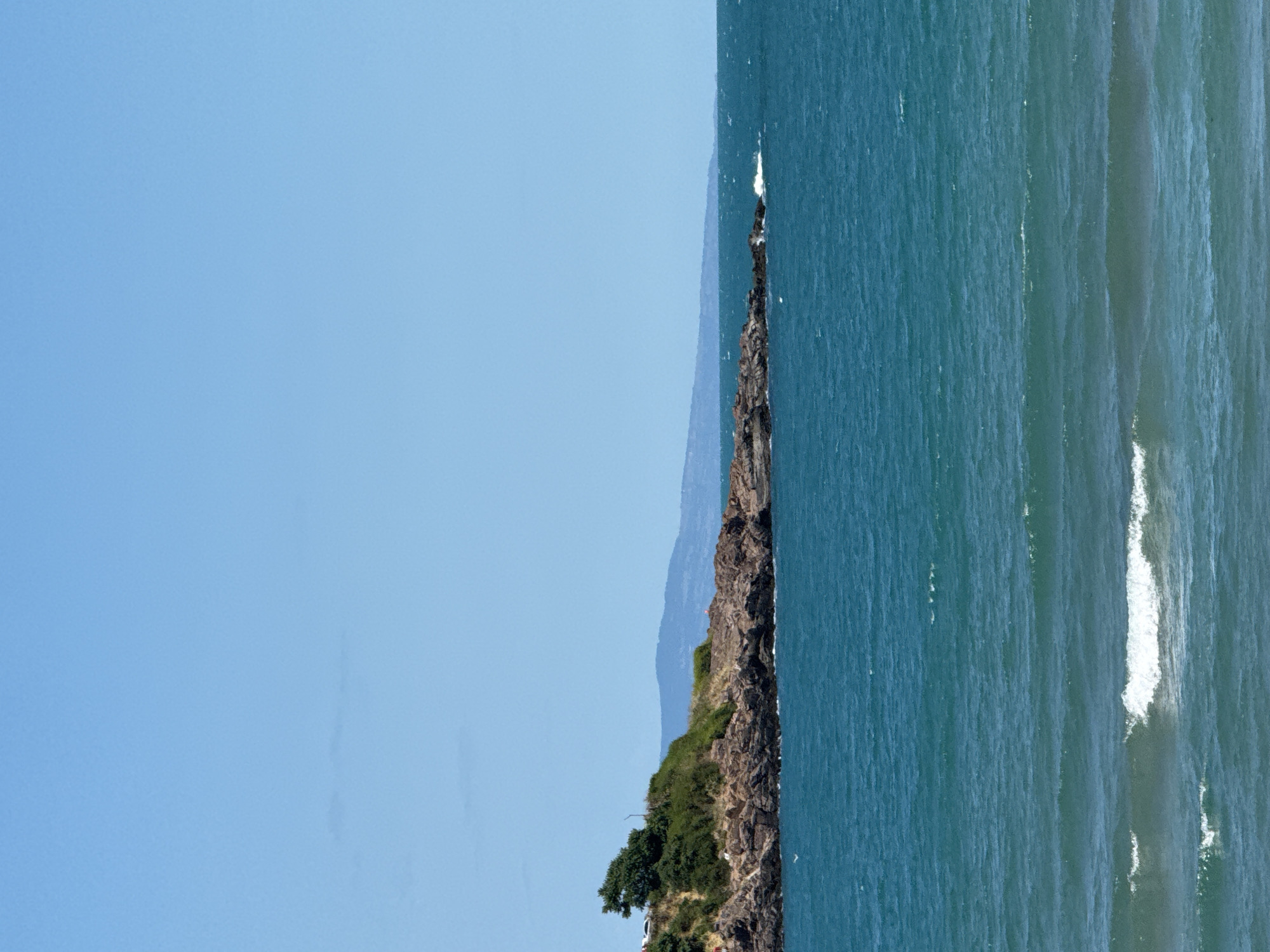 The image depicts a rocky coastline with turquoise water and a clear blue sky. A small island or peninsula jutting out into the sea is covered with green vegetation and rocks. In the distance, a hazy mountain range is visible on the horizon. Small white-capped waves are breaking against the shore. The scene is likely from Sozopol, Bulgaria, a coastal town known for its picturesque landscapes. No people are visible in the image.