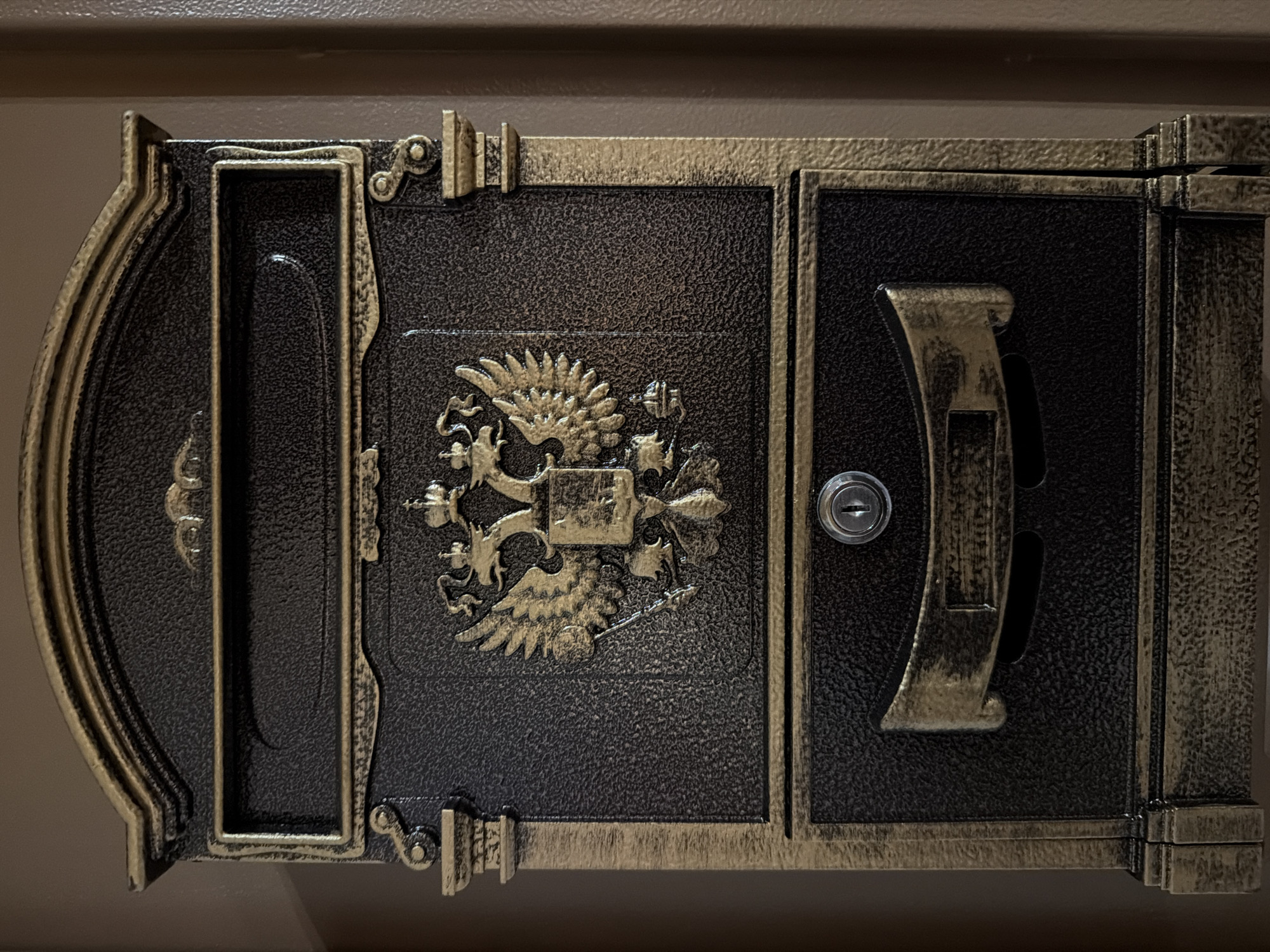 A close-up shot presents a dark, ornate metal door or panel featuring a prominently displayed double-headed eagle emblem, a symbol often associated with Russia. The metalwork exhibits a combination of dark and golden hues, contributing to an aged, potentially antique, appearance. A keyhole is visible on the right side of the panel. The image suggests a Russian cultural or historical connection, but without additional context or information, it is not possible to confirm the location in Krasnoyarsk or any other specific details about the scene.