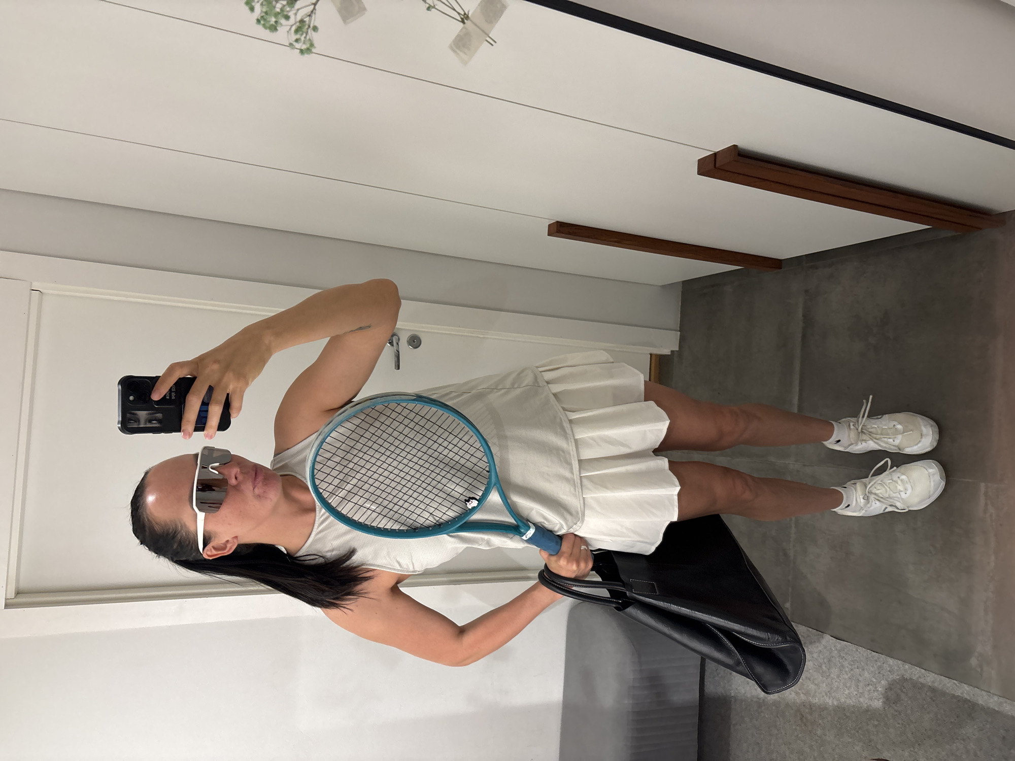 A woman is taking a selfie in a room with white walls. She is wearing a white tennis dress and white sneakers and sunglasses, and holding a tennis racquet and a black bag. She is holding a black smartphone. The location is not identifiable.