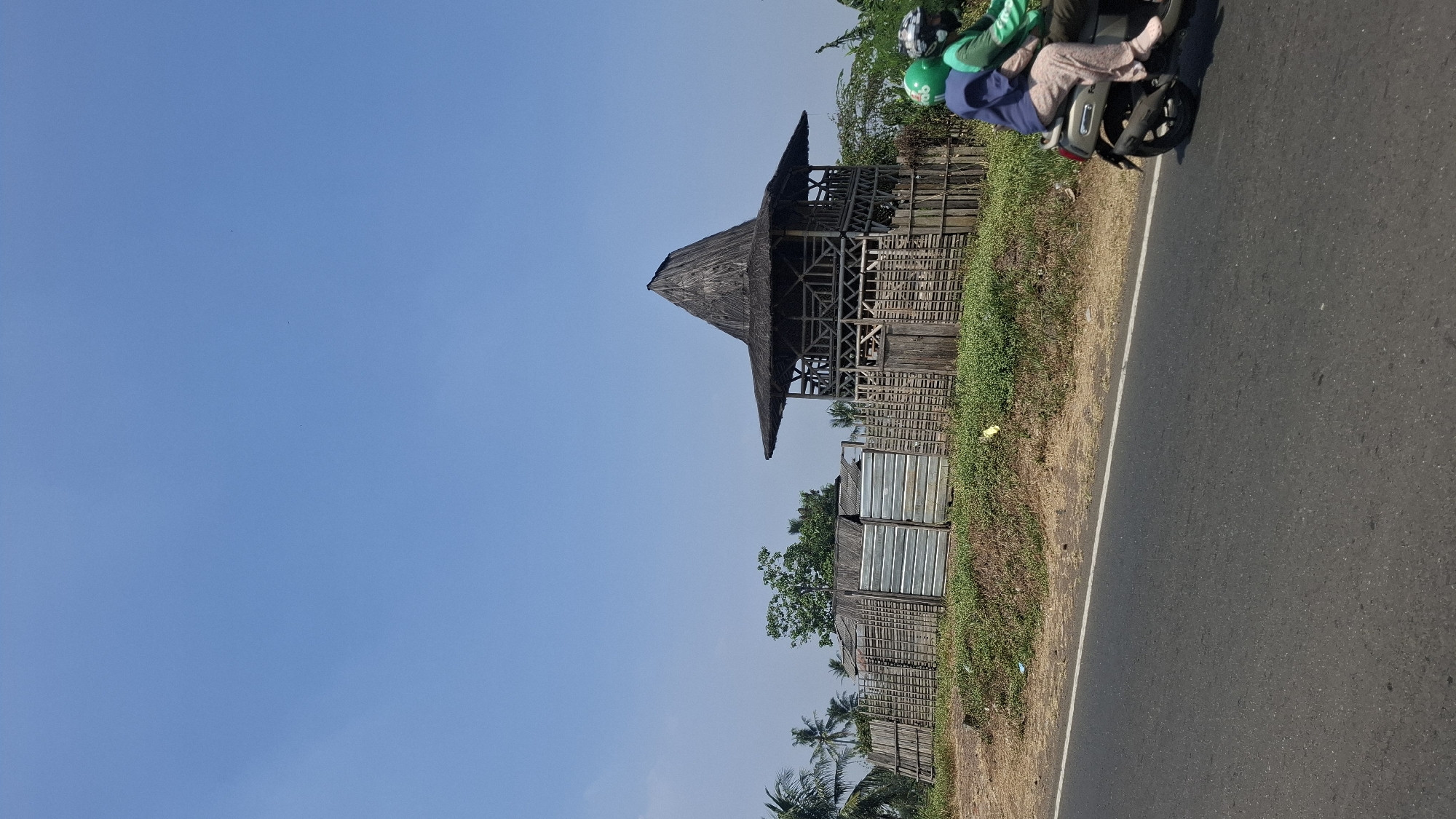 In Solokpandan, Indonesia, a motorcycle carrying two individuals passes by a traditional, conical-roofed structure with wooden lattice walls, possibly a gazebo or pavilion. The structure is situated behind a bamboo fence and a section of corrugated metal fencing. The scene is set against a clear blue sky, with the road in the foreground and vegetation, including palm trees, visible in the background. The people on the motorcycle are wearing helmets, one green and one dark, and the driver appears to be wearing a dark blue shirt and patterned pants, while the passenger wears a green jacket. The motorcycle is silver and black.