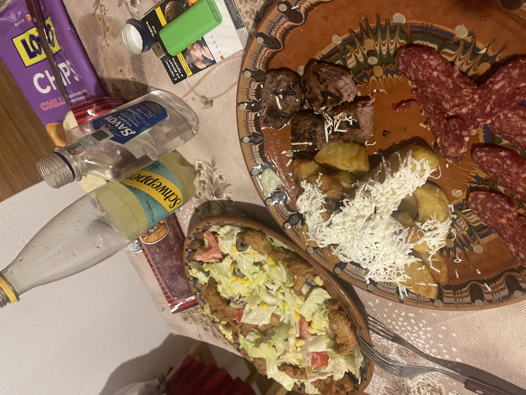 An indoor scene displays a meal laid out on a patterned tablecloth, featuring two main dishes and various accompaniments. On the right, a large, round, terracotta-colored plate with a colorful, ornate rim holds several pieces of cooked meat, possibly meatballs or grilled steak, some topped with grated cheese. Alongside these are fried potato wedges, also generously covered in grated cheese, and slices of cured sausage or salami.
To the left of the main plate, a smaller, oblong terracotta-colored bowl contains a salad composed of lettuce, chopped tomatoes, corn kernels, black olives, and pieces of fried, breaded food, likely chicken or fish.
Surrounding the food are several items: an empty clear plastic bottle with a blue label for "SAVOY VODKA 100% Pure EXCEPTIONAL QUALITY", and a partially filled glass bottle of "Schweppes BITTER LEMON". A purple bag of "LAYS CHIPS" (possibly "Chilli" flavor) is visible, as well as a packet of "KARE" cigarettes, priced at "€3.40 LEBGRPA", featuring a health warning in Bulgarian ("С тютюно на децат и прияте", "Помощ за отказ тютюно"). A green lighter and a white bottle cap are also on the table. Two forks with wooden handles are placed near the smaller salad bowl. The setting suggests a casual meal, possibly in the evening due to the indoor lighting.
