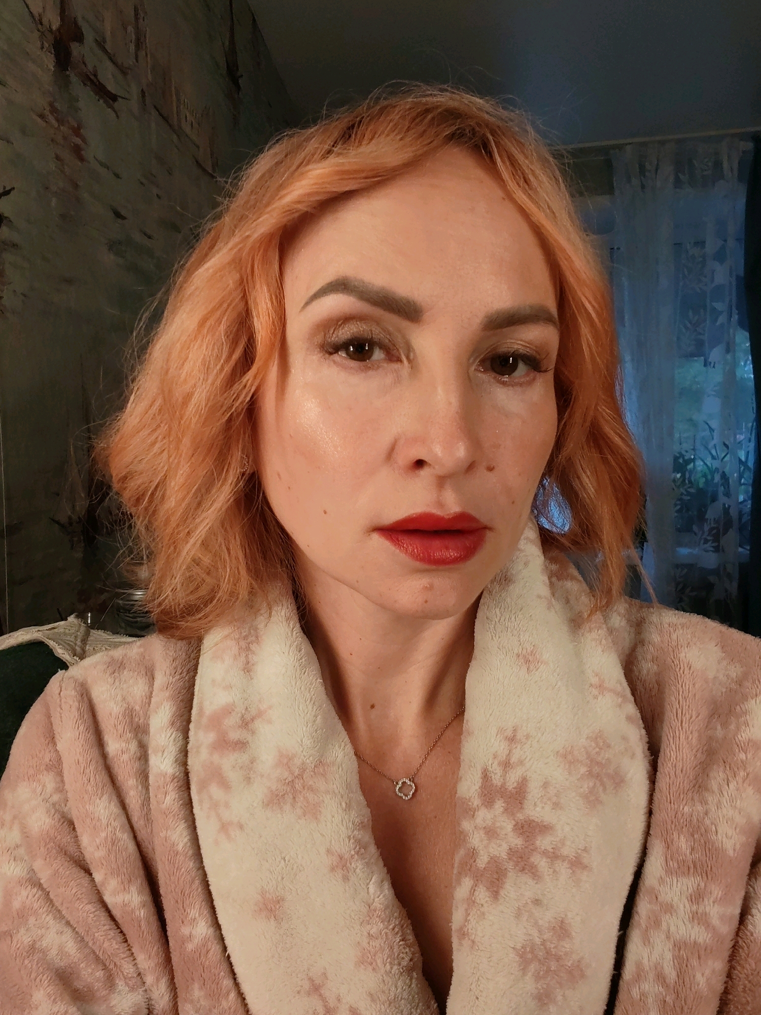 A close-up, waist-up portrait shows a Caucasian woman with shoulder-length, wavy, reddish-blonde hair. She is wearing a soft, pink bathrobe with a white fuzzy collar and a pattern of pinkish-red splotches. A delicate gold necklace with a small, four-leaf clover-shaped pendant adorns her neck. Her makeup includes defined eyebrows, eyeshadow, and bright red lipstick. The background is out of focus, but appears to be an indoor setting with patterned wallpaper to the left and sheer white curtains to the right. The lighting suggests it is daytime. There is no identifiable location in Chaikovsky, Russia.