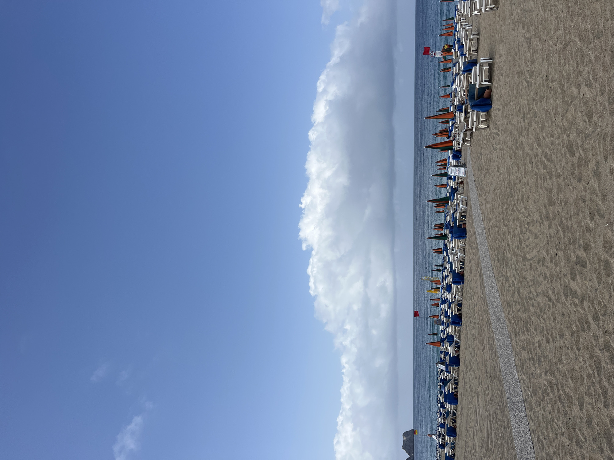 The image depicts a beach scene in Alanya, Turkey. The view is oriented parallel to the shoreline, with the sea on the right. A long stretch of sandy beach fills the lower right portion of the frame, populated by rows of white beach chairs and colorful sun umbrellas, including orange, green, and blue variants. A path or demarcation runs along the back of the beach seating. The Mediterranean Sea is visible as a thin band, bordering the beach. The sky occupies the majority of the left side of the image, transitioning from clear blue to a large formation of cumulus clouds. At the bottom right, a small rocky promontory extends slightly into the sea, marked with a lifeguard post, signified by two red flags.