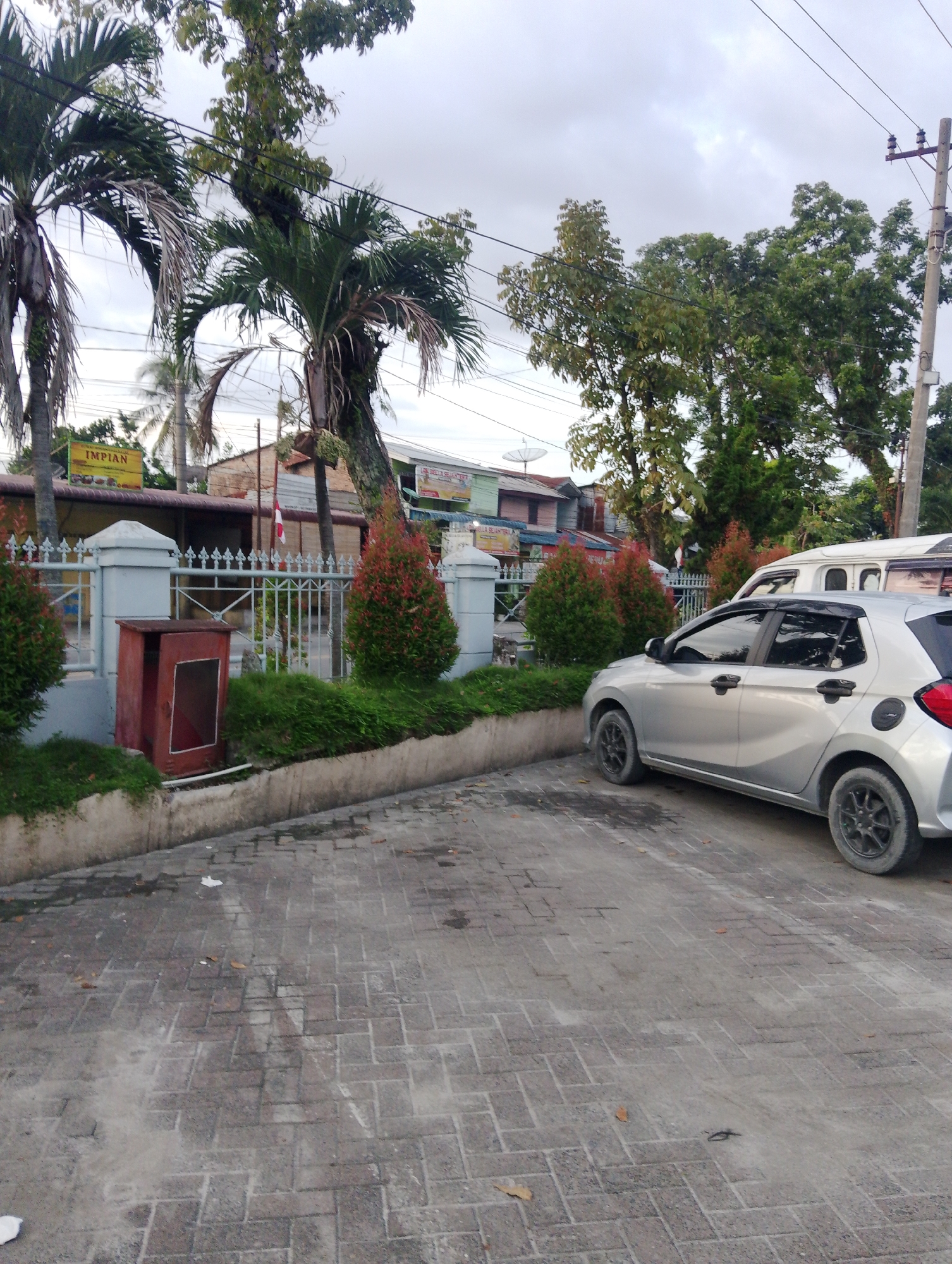 The image depicts an outdoor scene in the city of Pematang Siantar, Indonesia. In the foreground, a silver car is parked on a paved surface. The car has black trim around the windows and black rims on its wheels. To the left of the car, there is a white van. In the background, there is a white metal fence with decorative tops, behind which are some small, red-leafed bushes and a red fire extinguisher box. Further back, palm trees and other foliage are visible, along with buildings that appear to be shops or residences. A yellow sign with the word "IMPIAN" is also visible on one of the buildings. The sky is overcast.