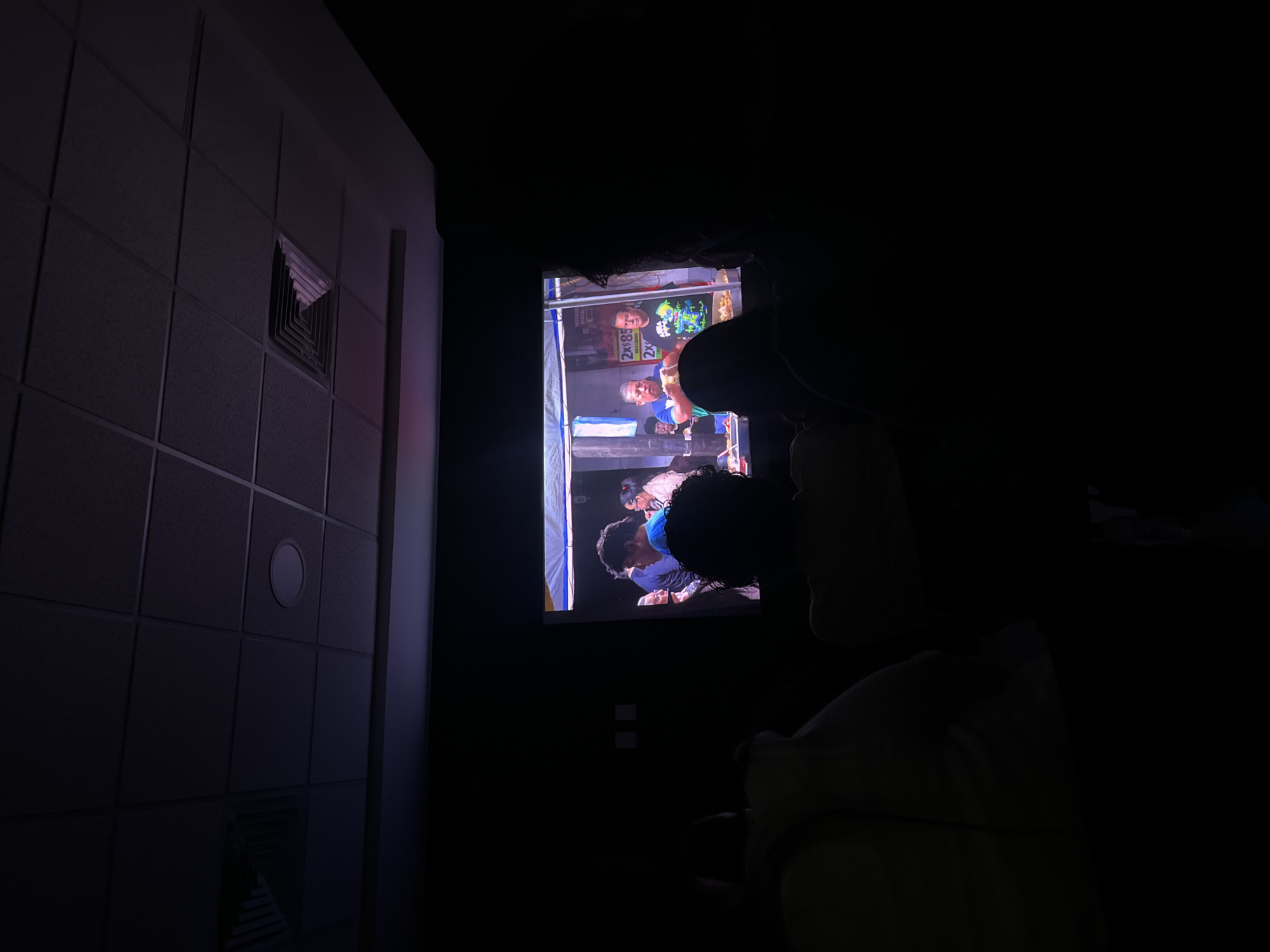The image shows a dimly lit room. On the left is a textured wall, possibly tiled, with what appears to be a ventilation grate near the top. A screen in the center is displaying a scene with people, presumably a video or a photograph. A figure is partially silhouetted in the foreground, possibly someone watching the screen. The location is unspecified in the image.