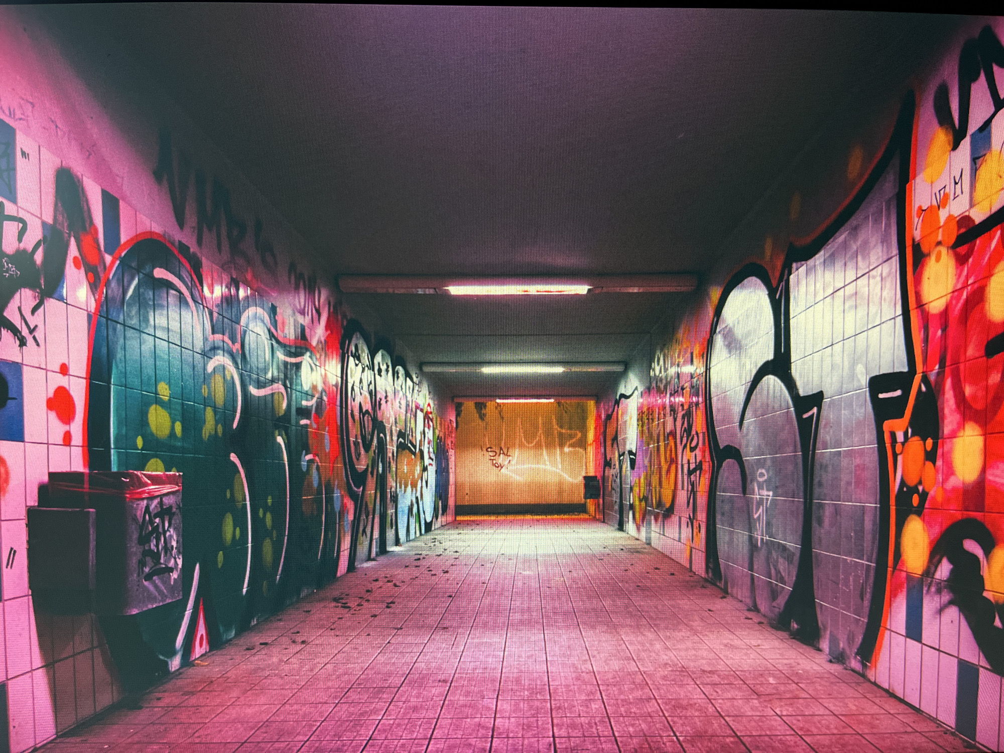 A dimly lit, graffiti-covered pedestrian underpass stretches into the distance. The walls are plastered with vibrant and chaotic graffiti in various colors, including red, yellow, green, and black. The ceiling features two fluorescent light fixtures that cast a warm glow down the tunnel. The floor is paved with square tiles, also with a pinkish hue, and littered with small debris. On the left wall, near the foreground, is a trash receptacle. The far end of the underpass is illuminated by an orange glow. The scene is devoid of any people.