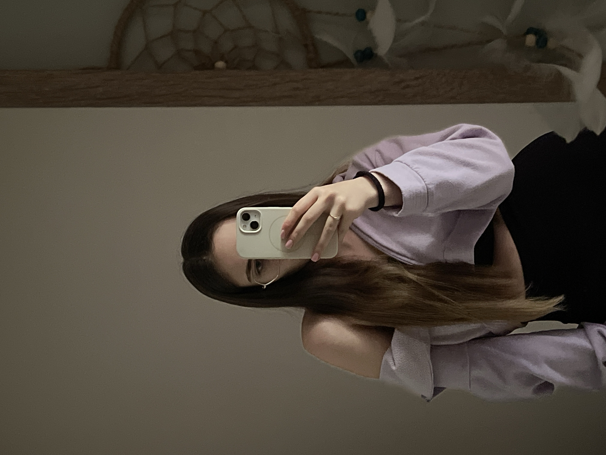 A woman is taking a selfie with a smartphone in front of a mirror. She is wearing a light purple sweatshirt and a black top. A dreamcatcher is visible in the mirror's reflection above a wooden shelf. The location is Unknown, Unknown.