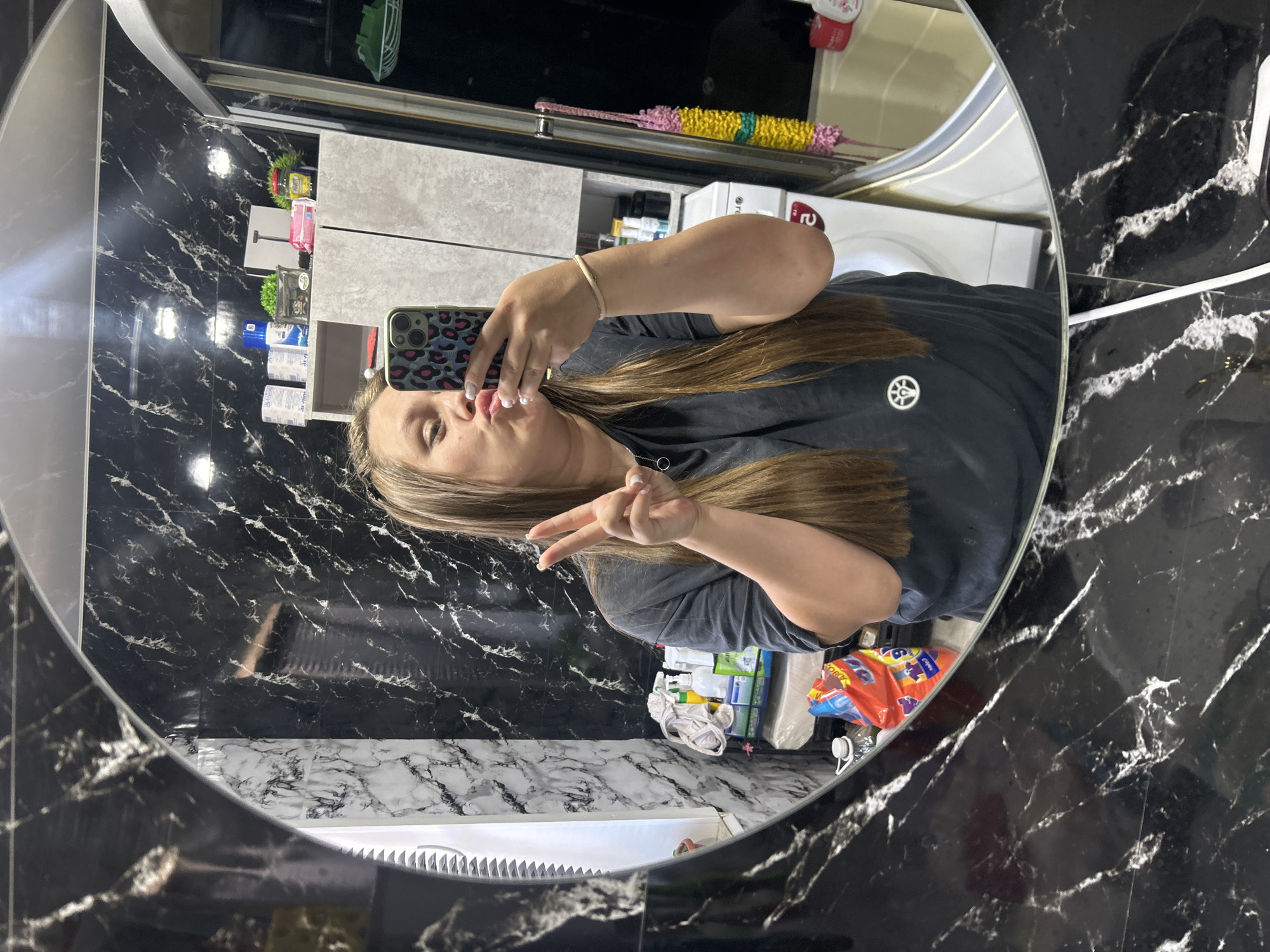 A young woman with long, brown hair is taking a selfie in a bathroom. She is wearing a dark grey t-shirt and is making a peace sign with her right hand while holding her phone in her left hand. Her lips are pursed as if she is about to kiss. The bathroom has black and white marbled walls. In the background, there are shelves with various cleaning supplies and toiletries. A washing machine is visible to the right of the shelves. The overall impression is a casual and personal moment captured in a domestic setting. The image does not provide any specific visual cues to indicate its location in Olkhovka, Russia, nor does it contain any elements that would suggest a forensic investigation is warranted.