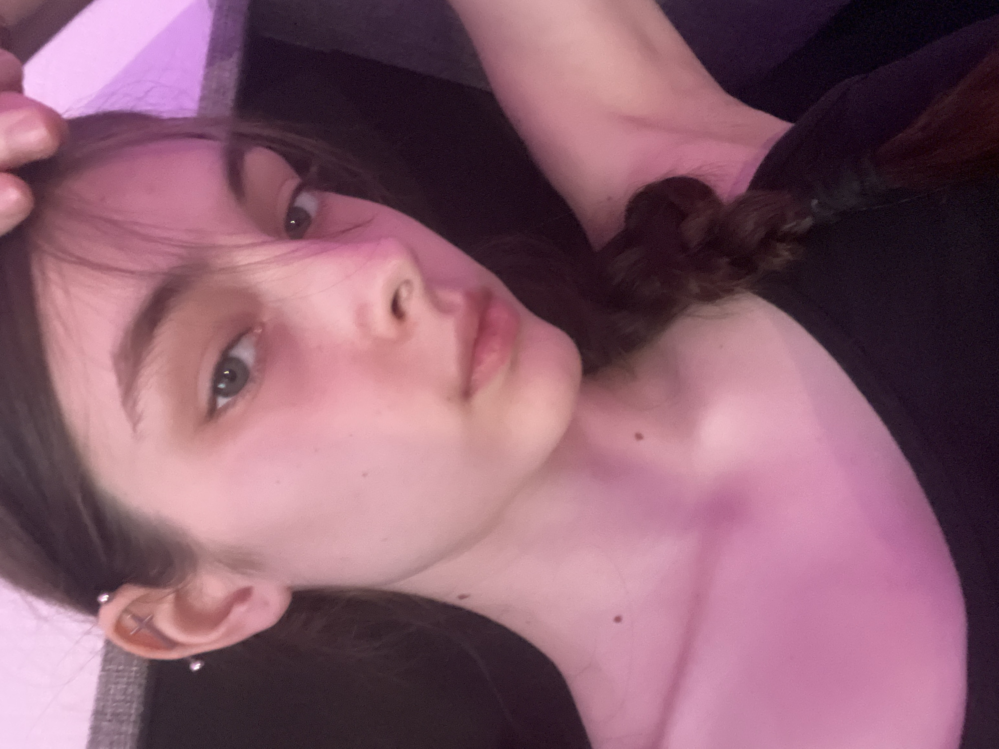 A close-up, rotated selfie of a young person with fair skin, light eyes, and brown hair with a braid visible. The individual is wearing a black shirt and has piercings in their left ear. The background is indistinct, with a glimpse of grey fabric on the left. The scene is bathed in a pinkish-purple light. The location is unknown and there is no evidence to suggest Saint Petersburg, Russia.