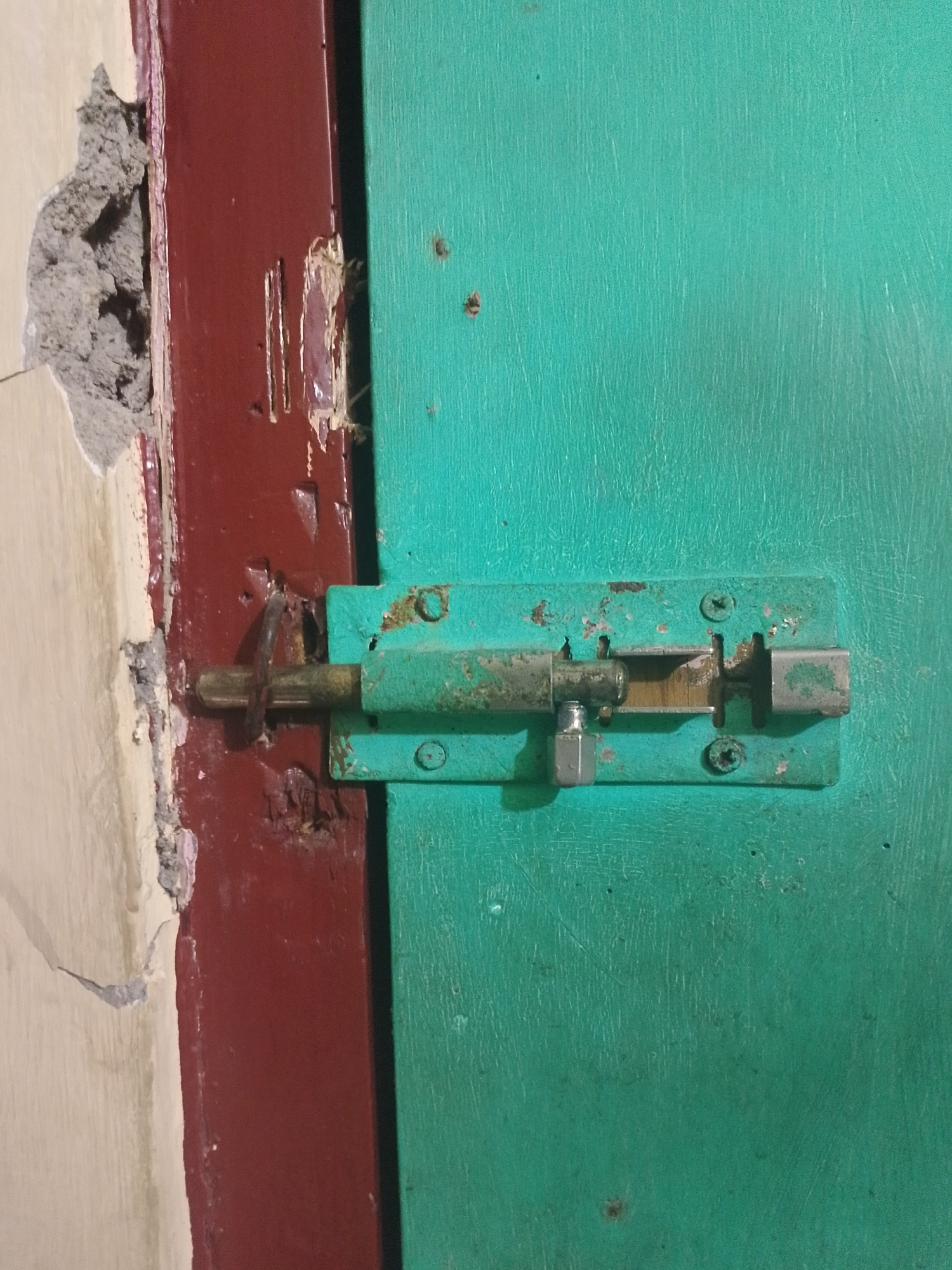 The image shows a close-up of a rusted green metal bolt lock on a teal-colored door. The lock is attached to the door with two visible screws. The bolt of the lock is extended and appears to be made of brass. To the left of the door, a dark red door frame is visible, with some peeling paint and exposed concrete or plaster. The wall to the far left is a light cream color with a damaged area showing rough gray material. The setting appears to be an interior doorway.