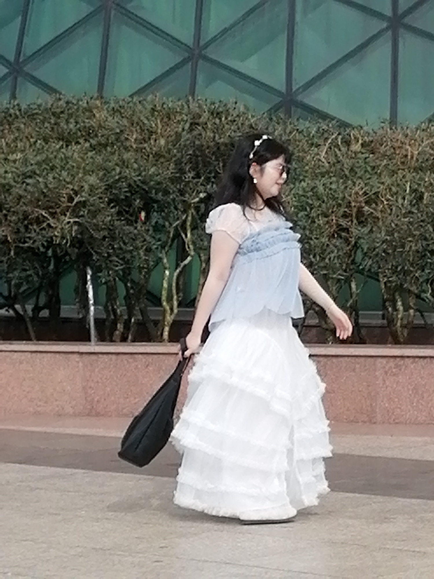 A young Asian woman walks in front of a dense green hedge in Da Lat, Vietnam. She wears a light blue ruffled top and a long, white layered skirt. A black bag is slung over her left shoulder. The woman has on round-rimmed glasses and a white hair accessory. In the background, a building with geometric windows is visible, and the ground is paved with concrete.