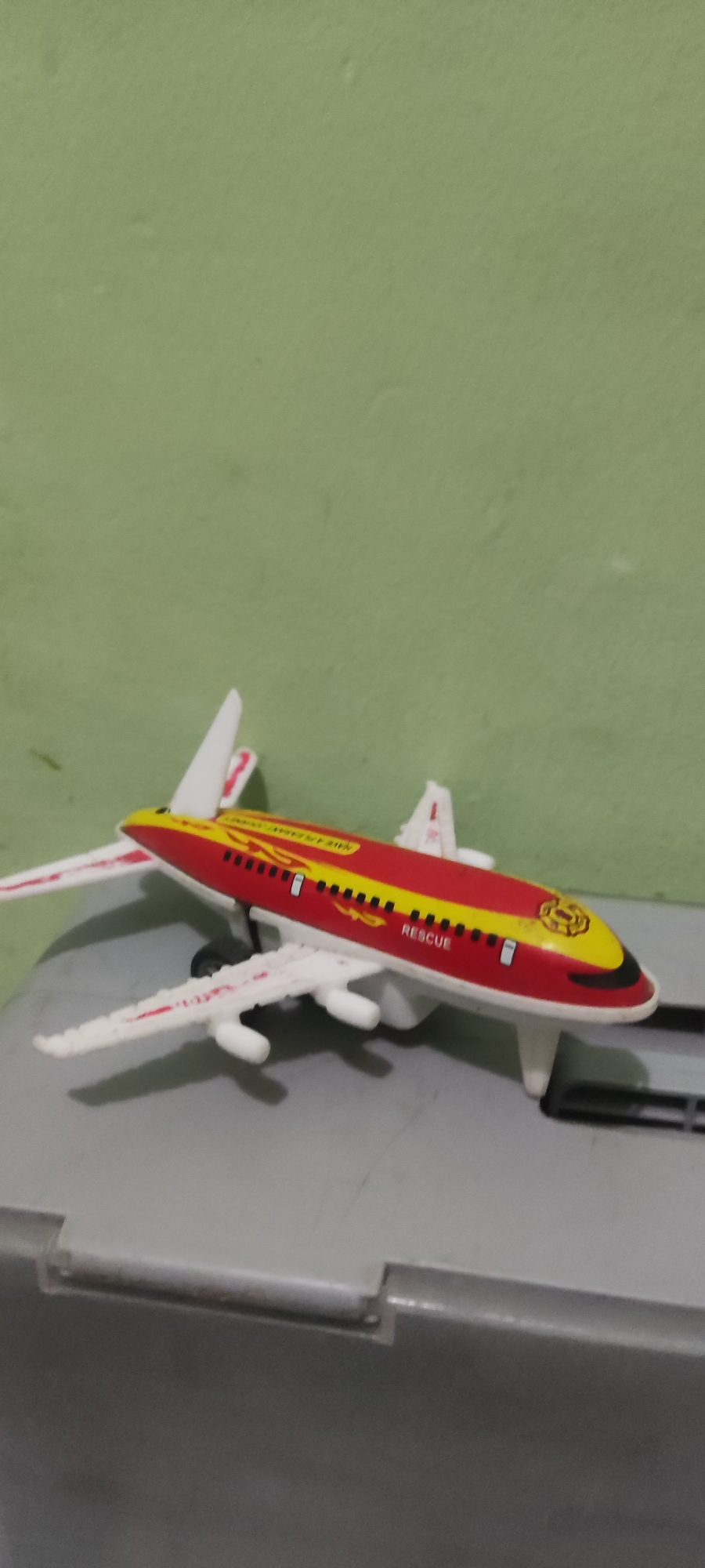 An overhead view captures a red and yellow toy airplane, identified as a rescue plane by the text on its fuselage. The airplane rests on a grey surface with black rails. The background consists of a light green wall. The image is a still photograph with no discernible activity or people. The location is identified as Gebang, Indonesia.