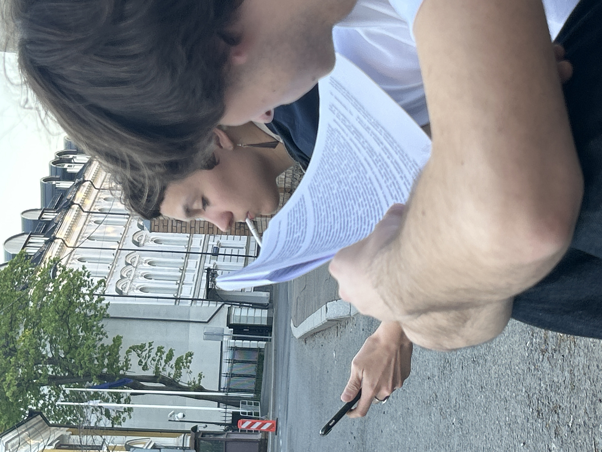 Two individuals are closely examining a sheet of paper filled with text. One individual holds a cigarette in their mouth. The other individual holds a smartphone in their right hand, with a wedding ring visible on their finger. The background shows a city street with European architecture.