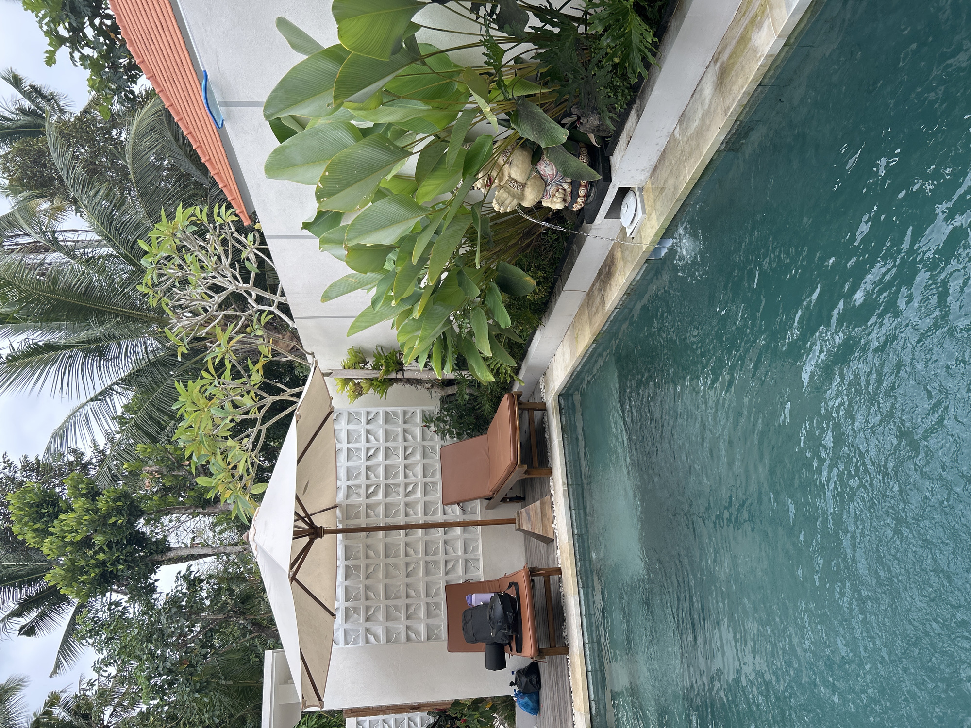 A medium shot captures an outdoor swimming pool area in Abiansemal, Indonesia. The pool, filled with clear turquoise water, is bordered by a light-colored tiled edge. To the left of the pool, two brown cushioned lounge chairs are placed on a wooden deck. The left chair has a black backpack and a water bottle on it, along with a rolled-up mat. To the right of the lounge chairs, a large white umbrella provides shade, supported by a wooden base. Behind the lounge chairs and umbrella is a white wall with a decorative, patterned texture. Lush green foliage, including palm fronds and broad-leafed plants, surrounds the area. On the right side of the pool, a small statue of a round figure is placed near the water's edge, with a stream of water flowing from it into the pool. The background is filled with more trees and vegetation, and a portion of a white building with an orange tiled roof is visible at the top right. The lighting suggests an overcast day.