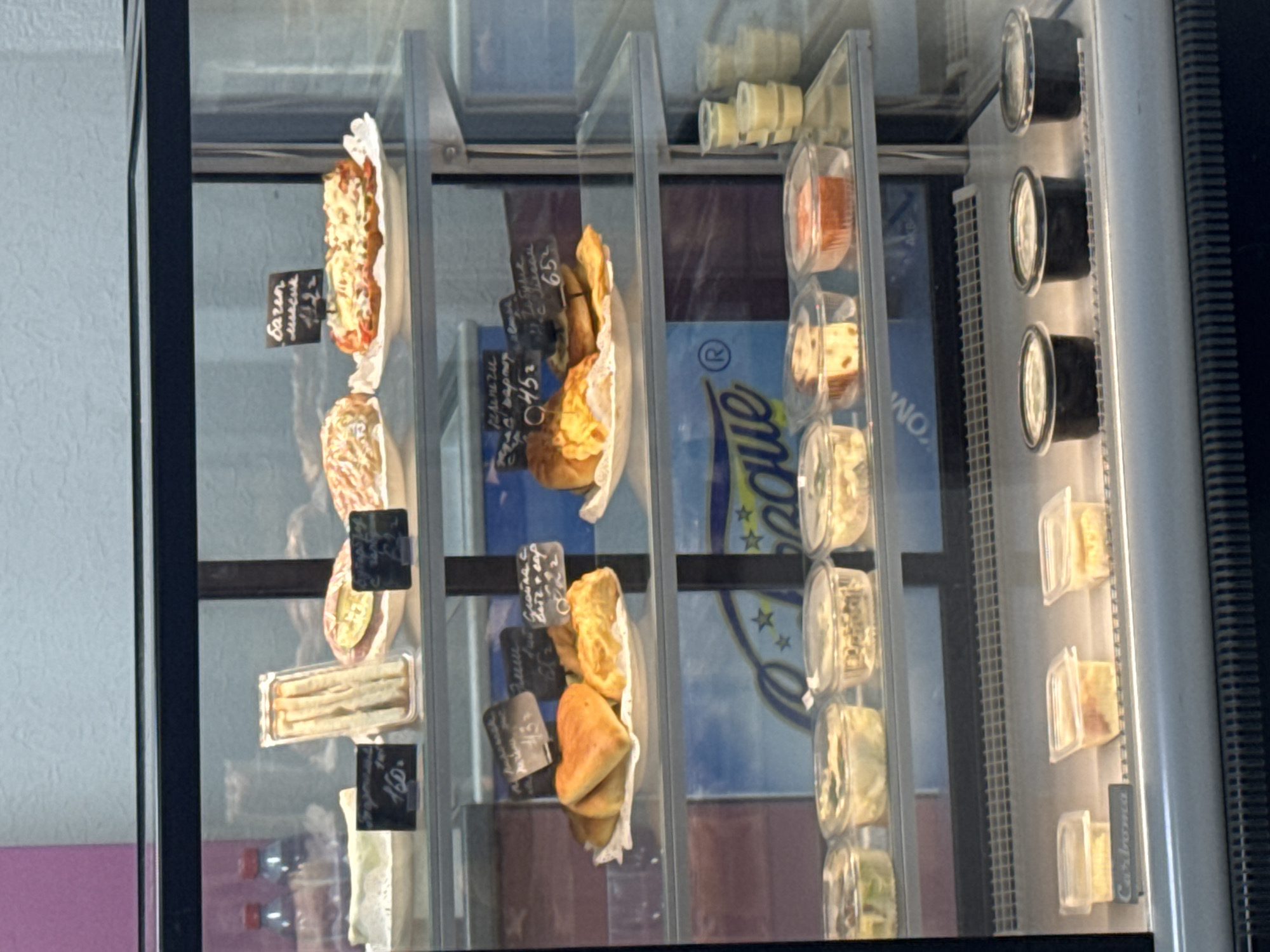 The image shows a refrigerated display case, likely in a food establishment. The case has multiple shelves containing various food items, including sandwiches, pastries, and pre-packaged meals or salads. Small black signs with white lettering are visible next to some items, presumably indicating the name and/or price of the food. In the background, through the glass of the display case, part of a sports team logo is visible on the back wall. Based on the visible logo (partial "League" text and blue/yellow colors) and references found, this is likely a display case in a store or café in Perm, Russia.