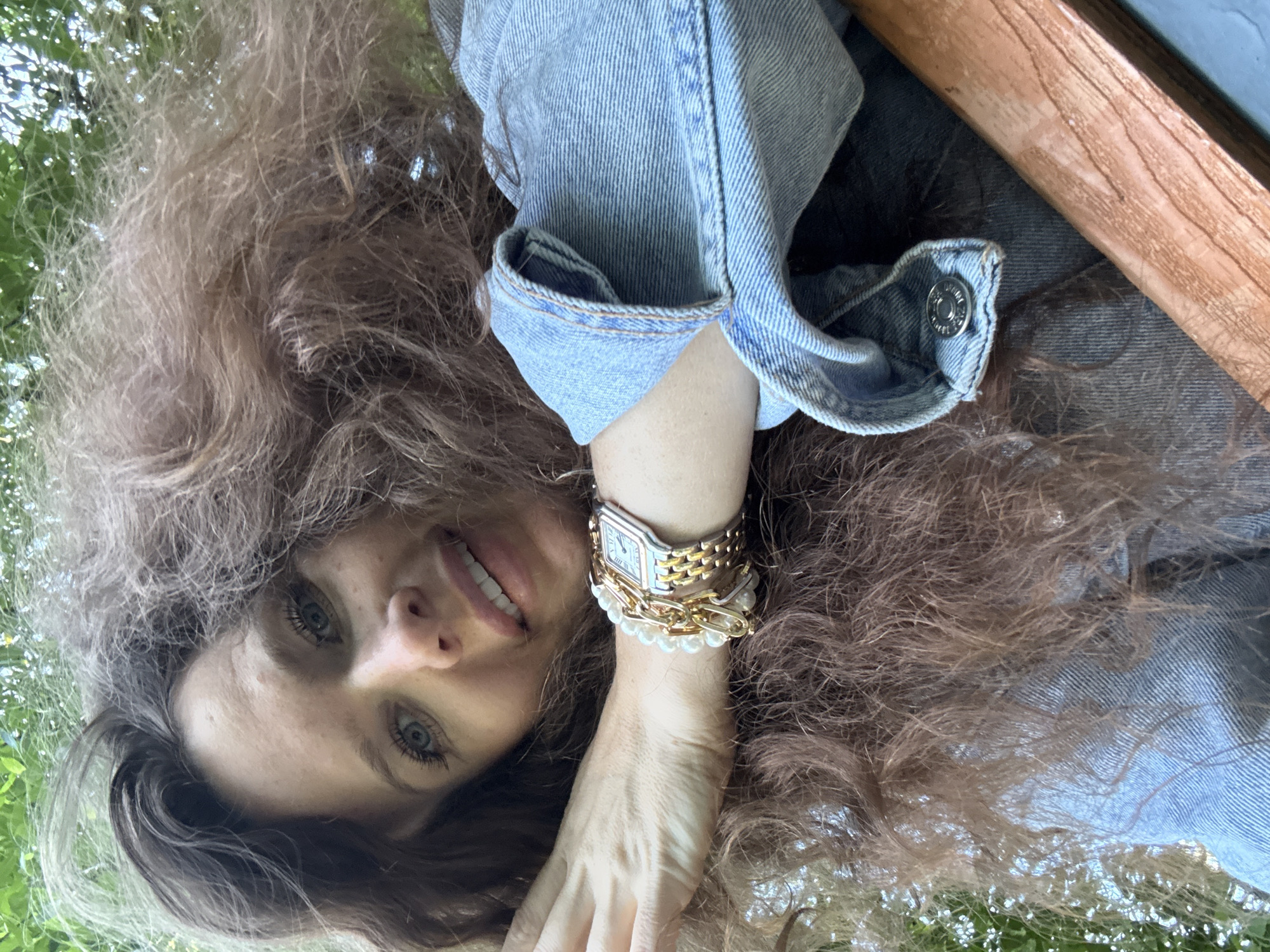 This is a close-up, overhead shot of a woman with long, curly, auburn hair. She is wearing a light blue denim jacket. Her face is tilted upwards, and she has bright blue eyes, long eyelashes, and a wide smile showing her teeth. She is wearing a watch on her left wrist along with several bracelets, including one with pearls. The background is blurred, with hints of green foliage and a wooden texture, suggesting an outdoor setting possibly in a park or garden. The lighting suggests it could be daytime, with natural light illuminating her face and hair. The overall mood appears cheerful and relaxed. There is no visible text in the image. The provided location context is Moscow, Russia, which adds a potential geographical element to the outdoor scene.