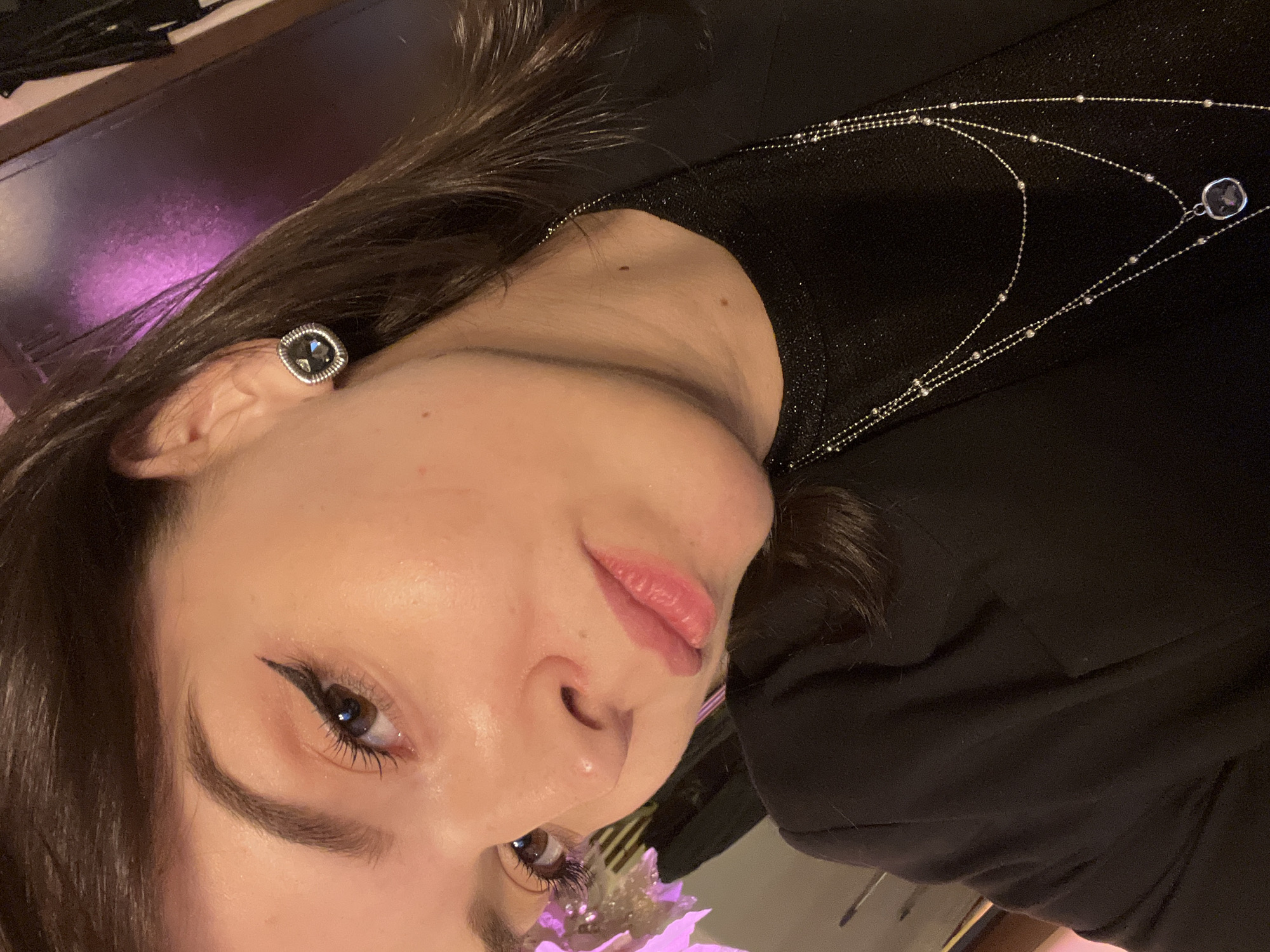 A close-up, slightly angled portrait of a woman with light skin, dark brown hair, and brown eyes. She is wearing a dark blazer and a delicate silver necklace. Her left ear is visible, adorned with a dark, square earring. She appears to be wearing eyeliner. The background is blurry, but suggests an indoor setting with subdued lighting and a hint of pink. The location in the city of Saint Petersburg, Russia is not discernible from the image.