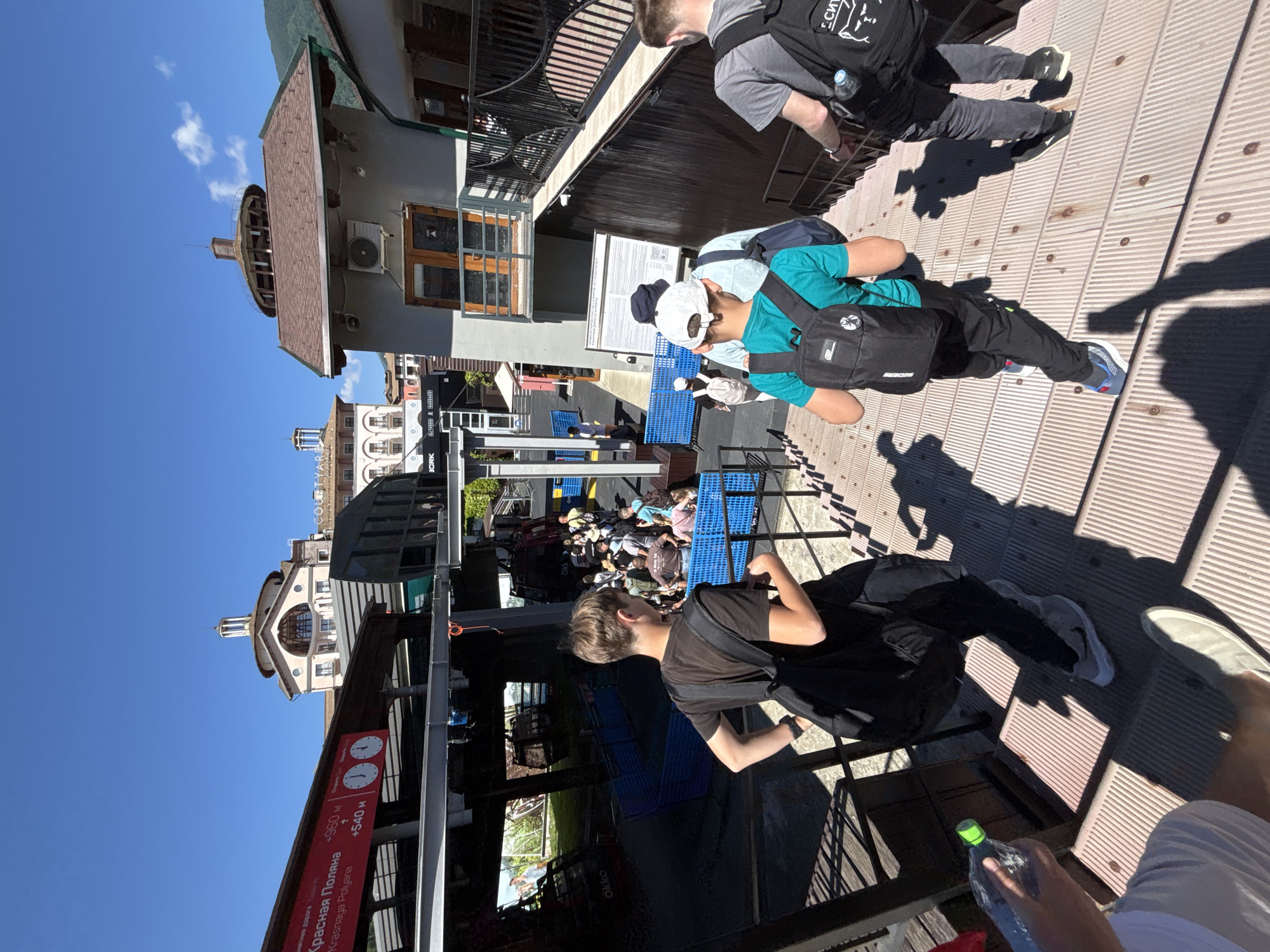 A group of people are ascending a wooden staircase under a clear blue sky. In the foreground, a young male wearing a black t-shirt and backpack is looking to his right. To his right, another young male in a teal t-shirt and backpack is walking up the stairs. Behind them, another person in a gray t-shirt and backpack is also walking up the stairs. In the background, a ski lift station is visible, with red and black gondolas parked. Signs in Russian are displayed, indicating information about the ski lift. Buildings with traditional architecture, including a domed structure, are visible in the distance. The overall environment suggests a tourist or recreational area in Estesadok, Russia, likely associated with a ski resort.