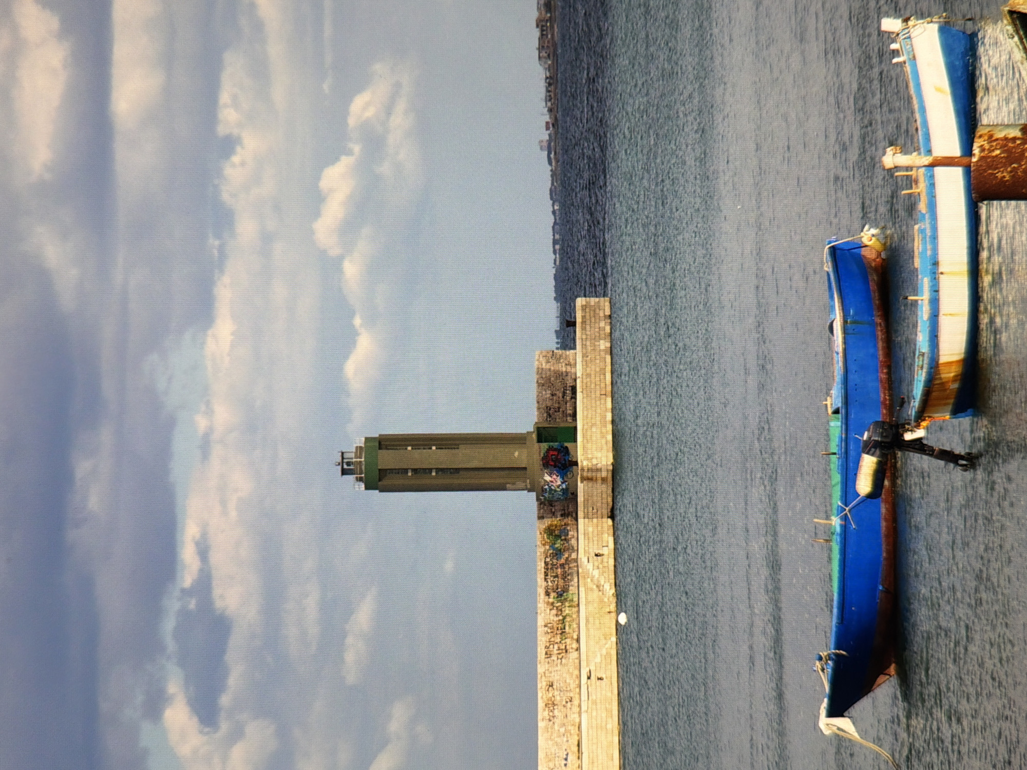 The image depicts a coastal scene with a tall, green lighthouse situated on a stone pier. The pier is adorned with graffiti. In the foreground, two boats are floating on the water, one predominantly blue and the other white and blue. The sky is filled with dramatic clouds. The water is calm. The scene is tranquil and appears to be a port or harbor area. The city of Kyiv, Ukraine is not depicted in this image; the setting appears to be a coastal town or city with a lighthouse and harbor.