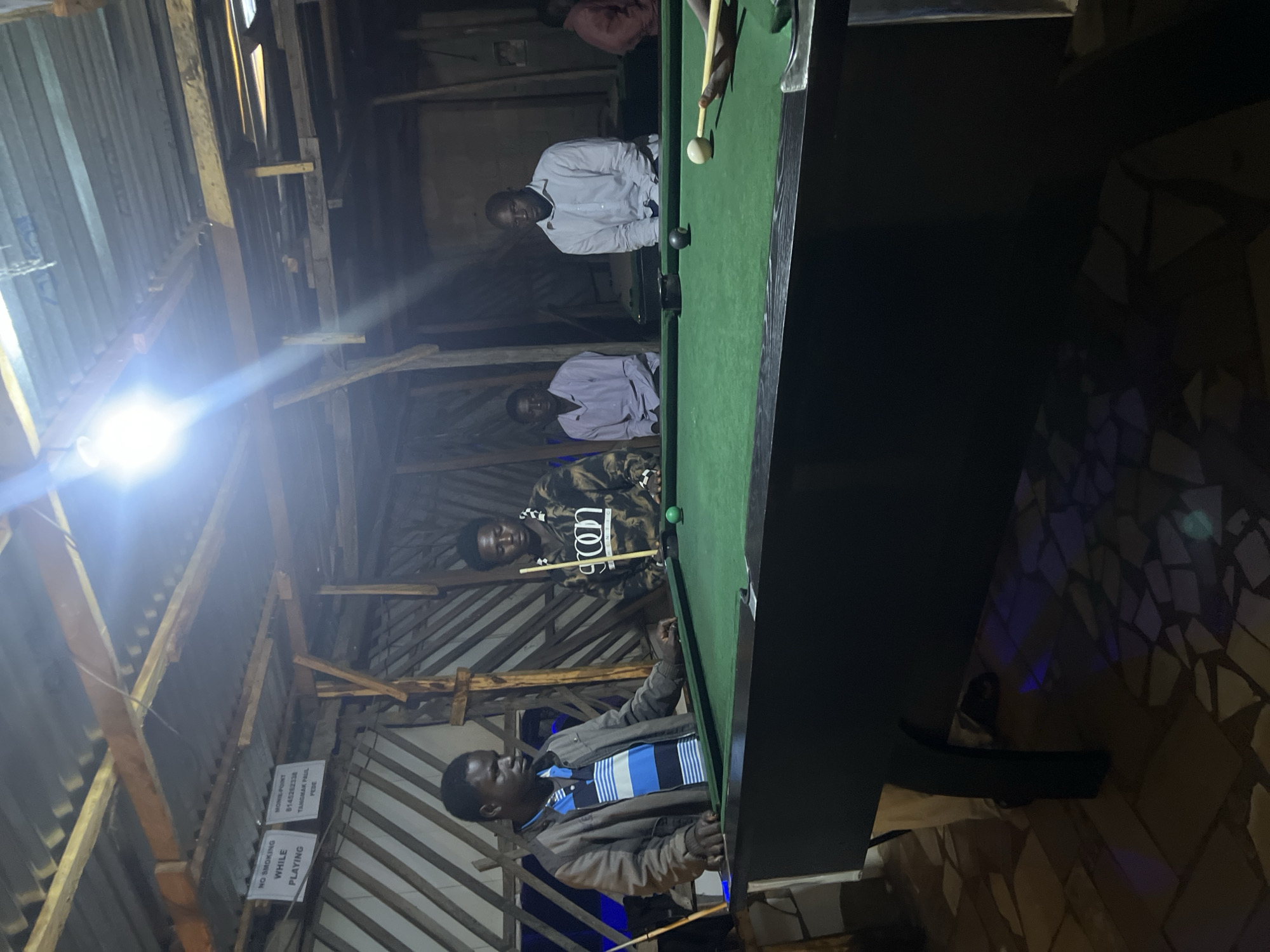 In Durumi, Nigeria, an indoor pool hall is depicted under dim lighting provided by a bright overhead bulb and possibly other ambient light sources. Four men are present around a green pool table. The man on the left, wearing a gray jacket over a striped blue and white shirt, stands with his hands on the table, appearing to be playing. To his right, a man in a tie-dye patterned hoodie holds a pool cue, aiming at a green ball on the table. Further to the right, two other men are seated, observing the game. One wears a light purple shirt, and the other a light blue button-down shirt. The room's structure is rustic, with a corrugated metal roof supported by exposed wooden beams and planks. Signs are visible on the wall, one reading "NO SMOKING WHILE PLAYING" and another with text that includes "MONIL POINT 6145262338" and "TANGMAI FAUL". The floor is tiled with a mosaic pattern of irregular stones. The overall atmosphere suggests a casual, recreational setting.