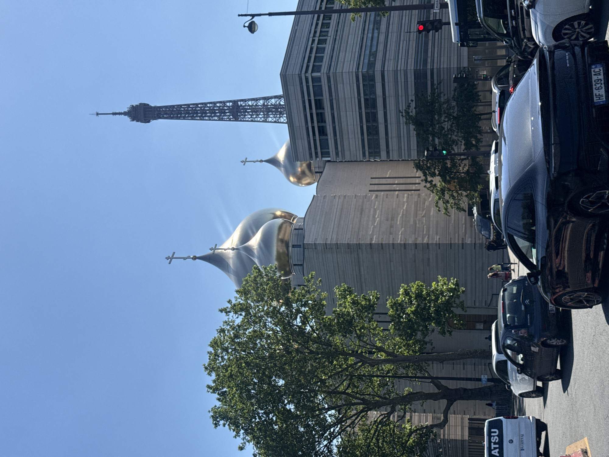 The image shows a street scene in Paris, France, featuring the Eiffel Tower in the background. In the foreground, the gold domes of the Holy Trinity Cathedral are visible. The cathedral is juxtaposed with a modern office building. A tree with green leaves partially obscures the view of the cathedral. The street is lined with parked cars, and people are walking on the sidewalk. A "MATSU" truck is also visible. Traffic lights are seen at the upper-right corner. The sky is blue and clear. Car license plate HF-639-AC is visible.