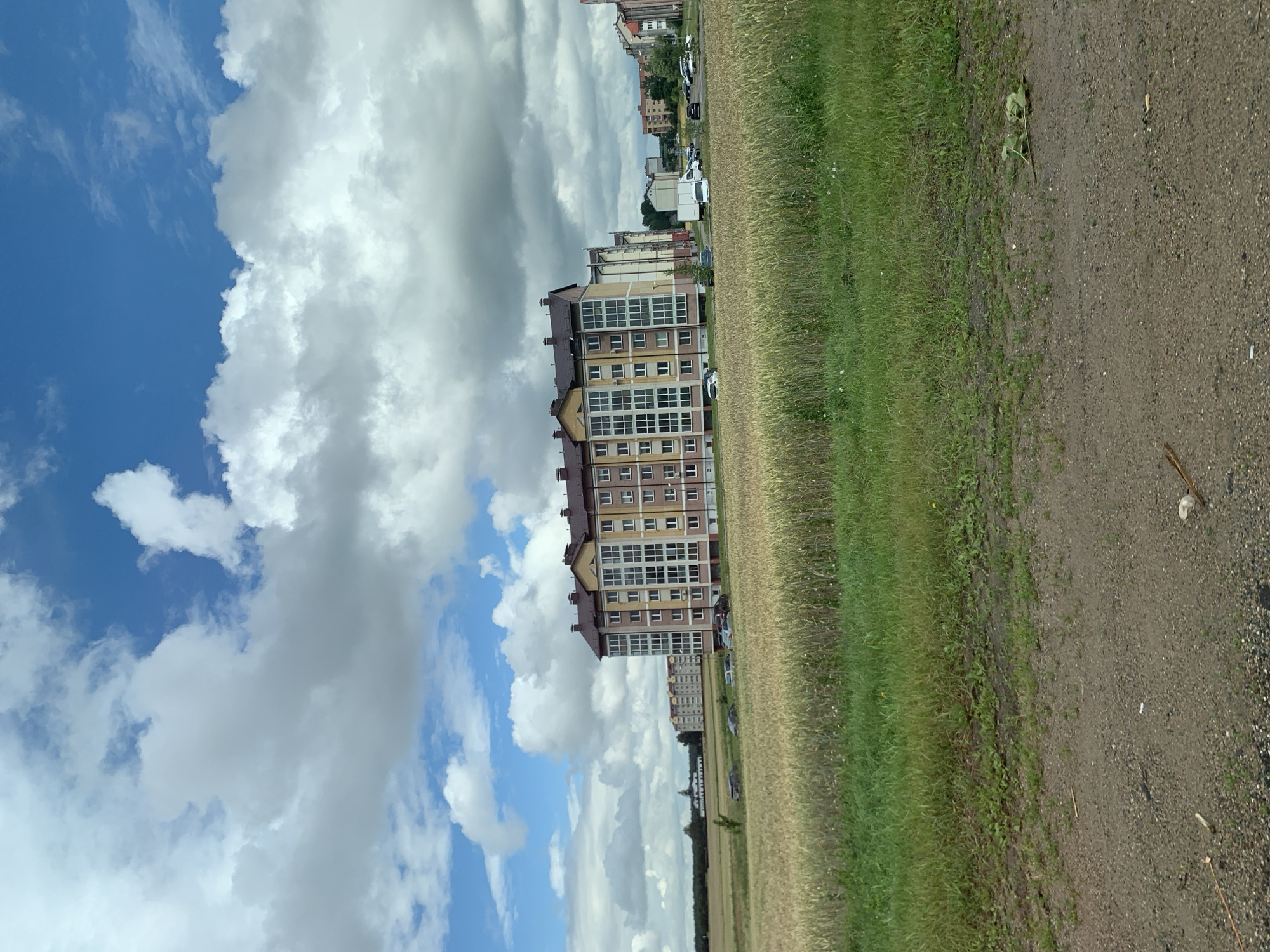 The image depicts a semi-urban or suburban landscape with a large, multi-story apartment building in the mid-ground. The building has a light yellow and pink facade with numerous windows and dark-colored roof structures. In front of the building, a field of mature wheat or similar grain is visible, with a strip of green grass and vegetation bordering a dirt or gravel path in the foreground. Several cars are parked in front of the apartment building. In the background, other residential buildings and some trees can be seen. The sky is partially filled with white and gray clouds against a blue sky, suggesting a daytime scene with mixed weather conditions. The overall environment suggests a residential area on the outskirts of a town or city, possibly in a rural or semi-rural setting within Нясвіжскі сельскі Савет, Belarus.