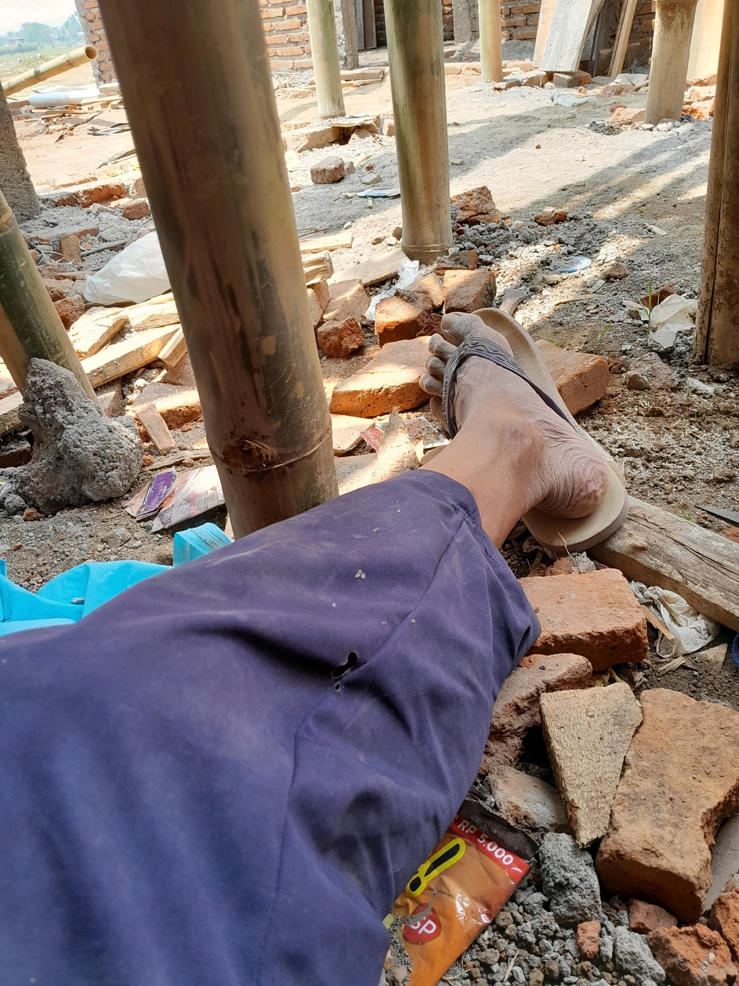The image depicts a construction site in Karangduren, Indonesia. A person's bare foot, clad in a flip-flop, rests on a pile of bricks. The person is wearing dark blue pants, which are torn in one area. The surrounding environment is characterized by scattered bricks, wood debris, and bamboo poles, indicating ongoing construction. The ground is covered in dirt and small stones. In the lower right portion of the frame, a discarded snack wrapper with Indonesian Rupiah currency (RP 5.000) is visible. The background reveals unfinished brick structures and more bamboo scaffolding, suggesting an incomplete building project. The lighting suggests it is daytime.