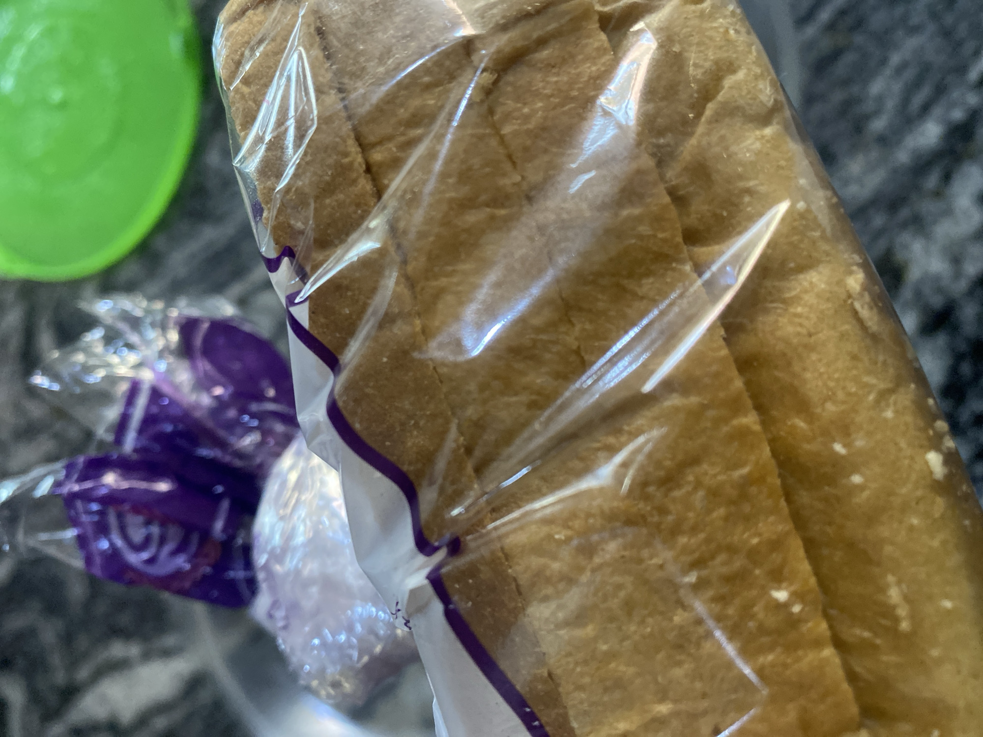 The image shows a close-up view of a loaf of bread, likely white bread, in clear plastic packaging. The bread is sliced and has a light brown crust. In the background, out of focus, are some purple-wrapped items, possibly candy or snacks, and a bright green lid of a container. The overall setting appears to be a kitchen or dining area, with a speckled countertop. The location of Bar Jirgi Summa, Nigeria, is not depicted or suggested by the visual elements in the image.