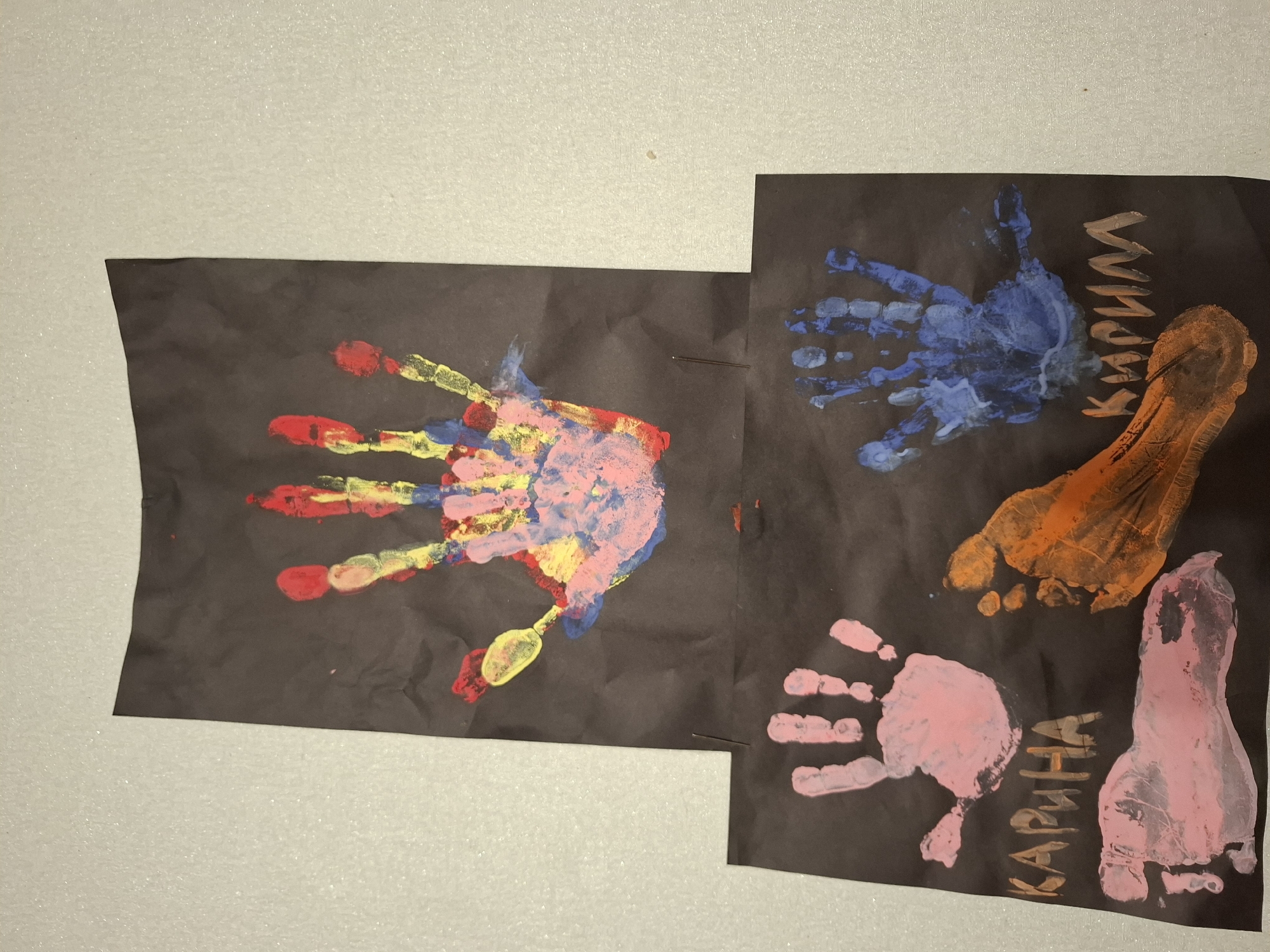 The image shows two pieces of black paper, each adorned with colorful hand and footprint art. The top piece of paper features a single handprint made with multiple colors, including red, yellow, pink, and blue. The bottom piece of paper displays several prints: a pink handprint on the left, a pink footprint below it, an orange footprint on the right, and a blue handprint above the orange footprint. There is also text written on the black paper. On the left side, beneath the pink handprint, the Cyrillic word "КАРИНА" is visible, spelled out in orange letters. On the right side, below the blue handprint and above the orange footprint, the Cyrillic word "КИРИЛЛ" is visible, spelled out in white letters. The papers are placed on a light-colored, textured surface, possibly a wall or a table. The scene is indoors, and the lighting suggests an artificial light source. There are no people visible in the image, but the hand and footprints imply the presence of children or adults who created the artwork. The location of Новлянка, Russia, is not evident from the image itself, as there are no identifiable landmarks or contextual clues.