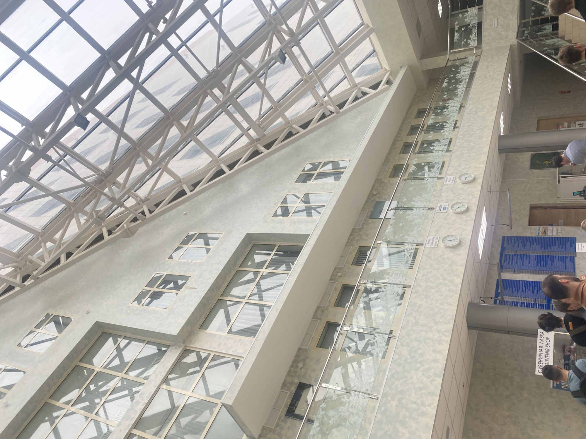 The image depicts the interior of a building, likely an airport, in Прикамский, Russia. A large glass roof supported by a white metal framework dominates the upper left portion of the image, allowing natural light to filter into the space. The building's walls are a light gray color and feature several rectangular windows with white frames, positioned in a somewhat staggered pattern.
A prominent feature is a tall, narrow glass wall or partition, potentially an elevator shaft or decorative structure. It runs vertically through the center of the image, adorned with a pattern of stylized white floral or leaf designs. Several clocks are mounted along this glass wall, presumably indicating different time zones.
On the right side of the image, a section of the building's interior is visible. There are walls, doors, and informational boards with blue lettering. Several people are present, though their faces are mostly obscured. The back of one person's head is clearly visible. There is also a sign in Russian that reads "СУВЕНИРНАЯ ЛАВКА/SOUVENIR SHOP".