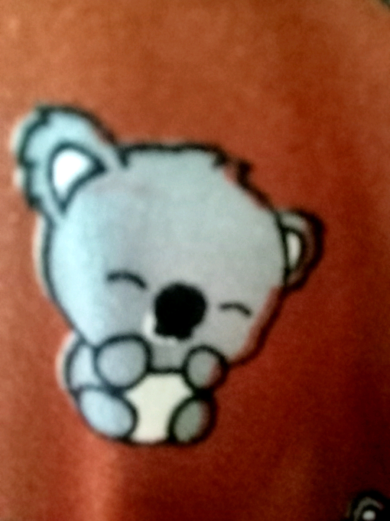 The image displays a cartoonish rendering of a koala, possibly an applique or sticker, on a textured, reddish-orange surface. The koala is predominantly light gray with a black nose, black curved lines for eyes, and a white belly. Its ears are large and rounded, and its limbs are depicted as simple, rounded shapes. The background is out of focus, showing a uniform, warm color. There is no discernible indication of Jalandhar, India, in the image.