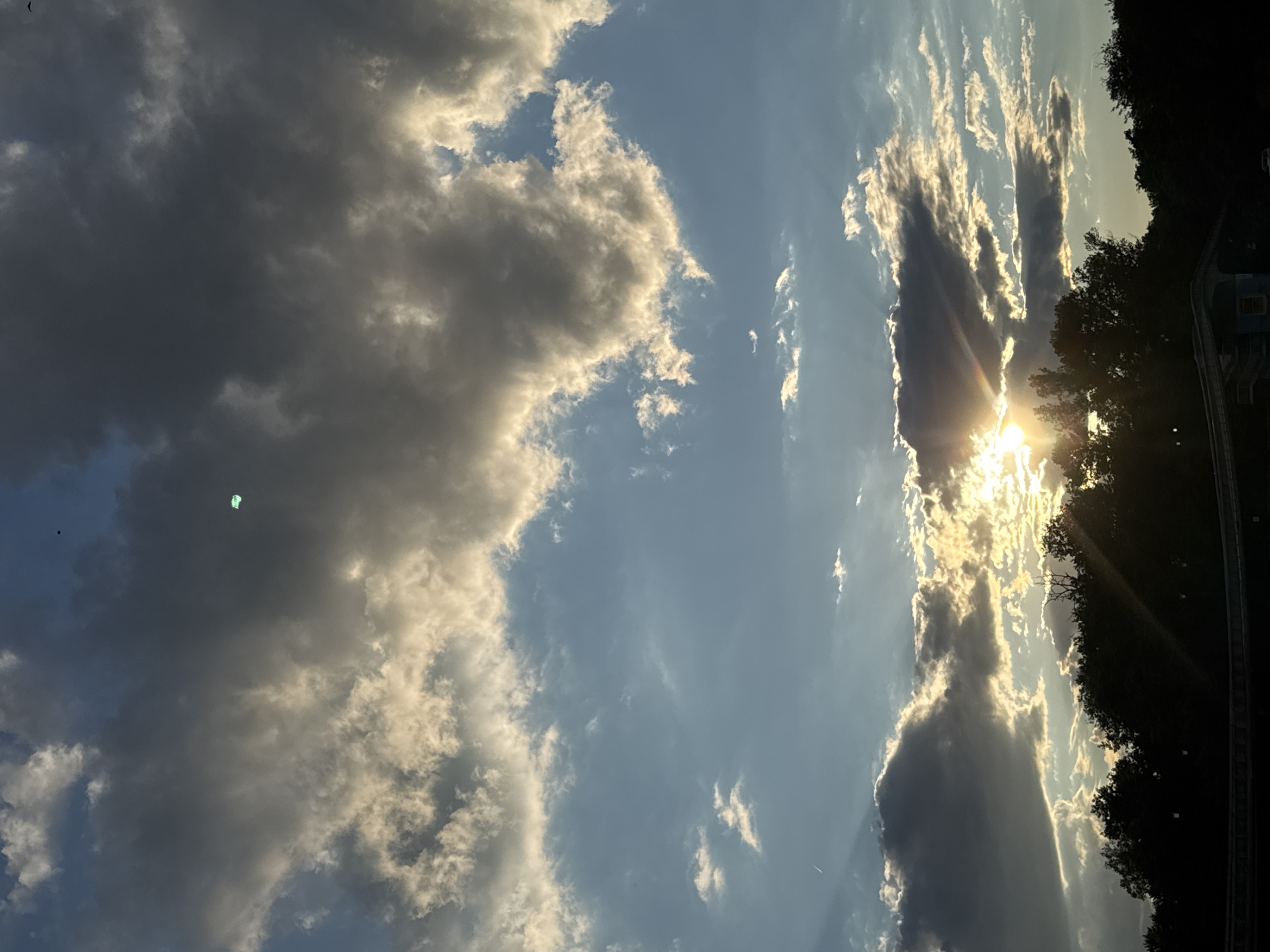The image depicts a dramatic sky with the sun setting behind silhouetted trees and foliage, casting light rays through dark, scattered clouds. The sun is partially obscured by a dark cloud formation on the lower right side of the frame. Wispy clouds are visible higher in the sky, catching the sunlight. A dark band, possibly representing a road or landscape feature, runs along the bottom of the image, with hints of structures and faint lights visible within it. The overall scene suggests an outdoor setting at dusk, potentially in a park or along a roadside within Plovdiv, Bulgaria, given the context.