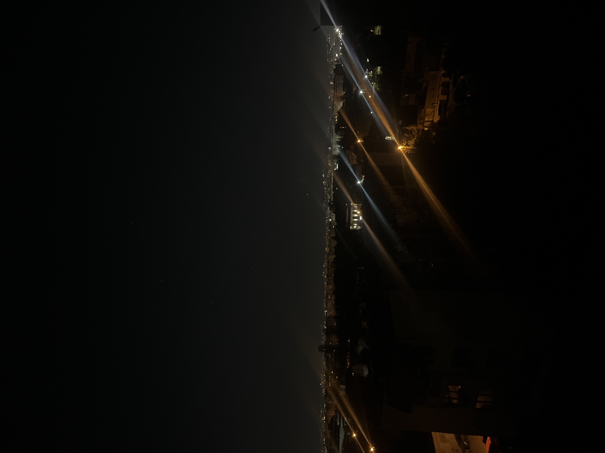 The image presents a nocturnal panoramic view of what appears to be a large urban or suburban area, identified as Sofia, Bulgaria, captured from an elevated perspective. The scene is predominantly dark, illuminated by numerous artificial light sources.
The upper portion of the frame is dominated by a deep black night sky, with a few extremely faint points of light possibly indicating distant stars or minor atmospheric phenomena. A noticeable atmospheric haze or light pollution subtly brightens the sky closer to the horizon line.
Running horizontally across the mid-section of the image is a vibrant, extended line of lights, representing the distant cityscape. These lights, likely from buildings, streetlights, and possibly vehicles, form a shimmering band that stretches into the hazy distance, marking the vastness of the urban development. The haze slightly blurs the individual light sources further away.
In the foreground, particularly towards the bottom-right, several brighter and closer artificial light sources are prominent. These lights exhibit distinct lens flares and streaks, characteristic of intense illumination against a dark backdrop. The immediate surroundings of these foreground lights are largely obscured in shadow, with only vague, dark outlines of structures or dense foliage barely discernible.
The overall scene conveys the quiet grandeur of a city at night. The time is clearly night, and the weather appears clear, although atmospheric light pollution is evident. No specific activity, identifiable individuals, or legible text are visible in the photograph.