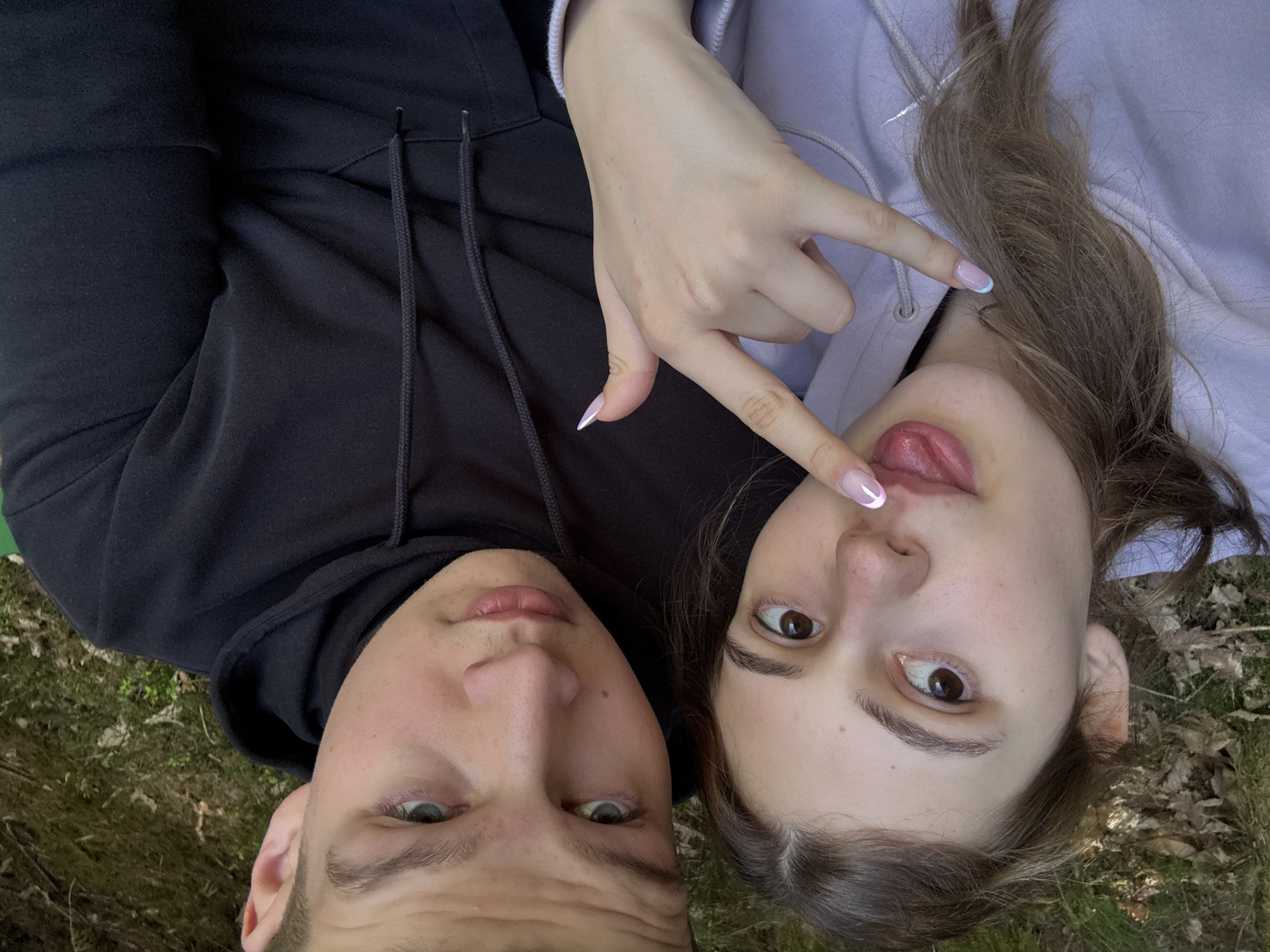 Two individuals are lying on what appears to be grass or foliage, possibly in an outdoor setting. One person, presumably male, is wearing a black hoodie. The other person, presumably female, is wearing a light purple hoodie and is sticking their tongue out while making the "hang loose" hand gesture. The image is taken from an overhead perspective, looking directly down at the subjects. The location is not discernible from the image. I do not have enough information to determine if it's in Forbach, Germany.