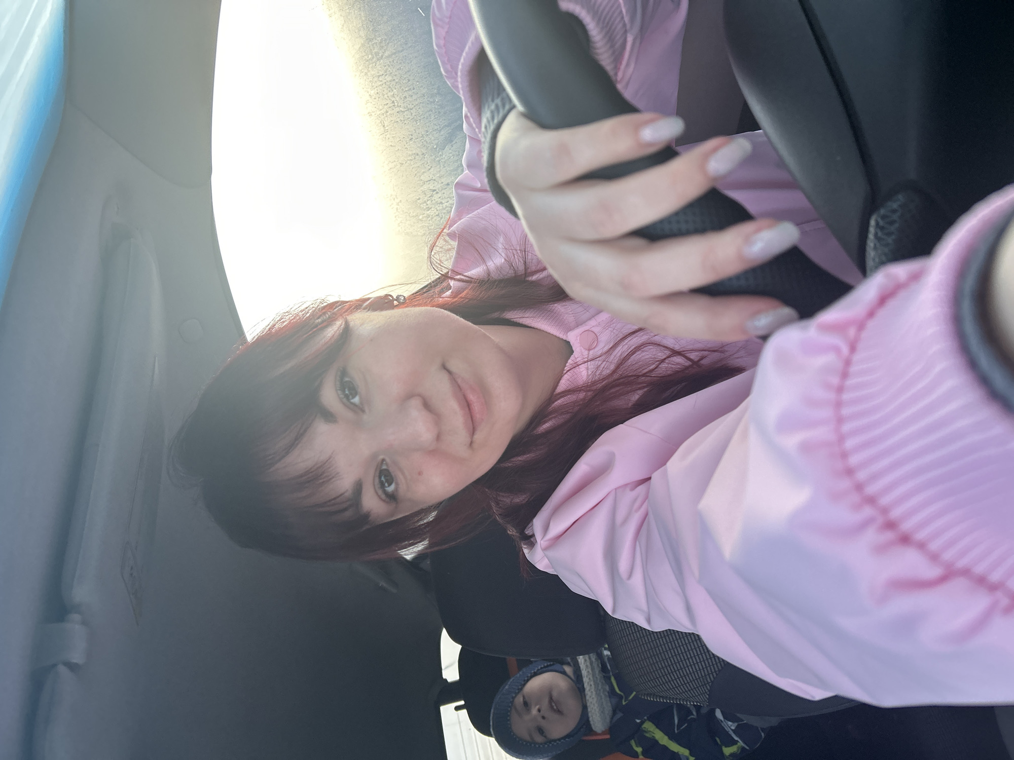 A woman with red hair is in a car, holding the steering wheel. She is wearing a pink jacket. A child in a blue hat and patterned jacket is in the background, likely in a car seat. The image appears to be taken from inside the car. The location is Фоки, Russia.