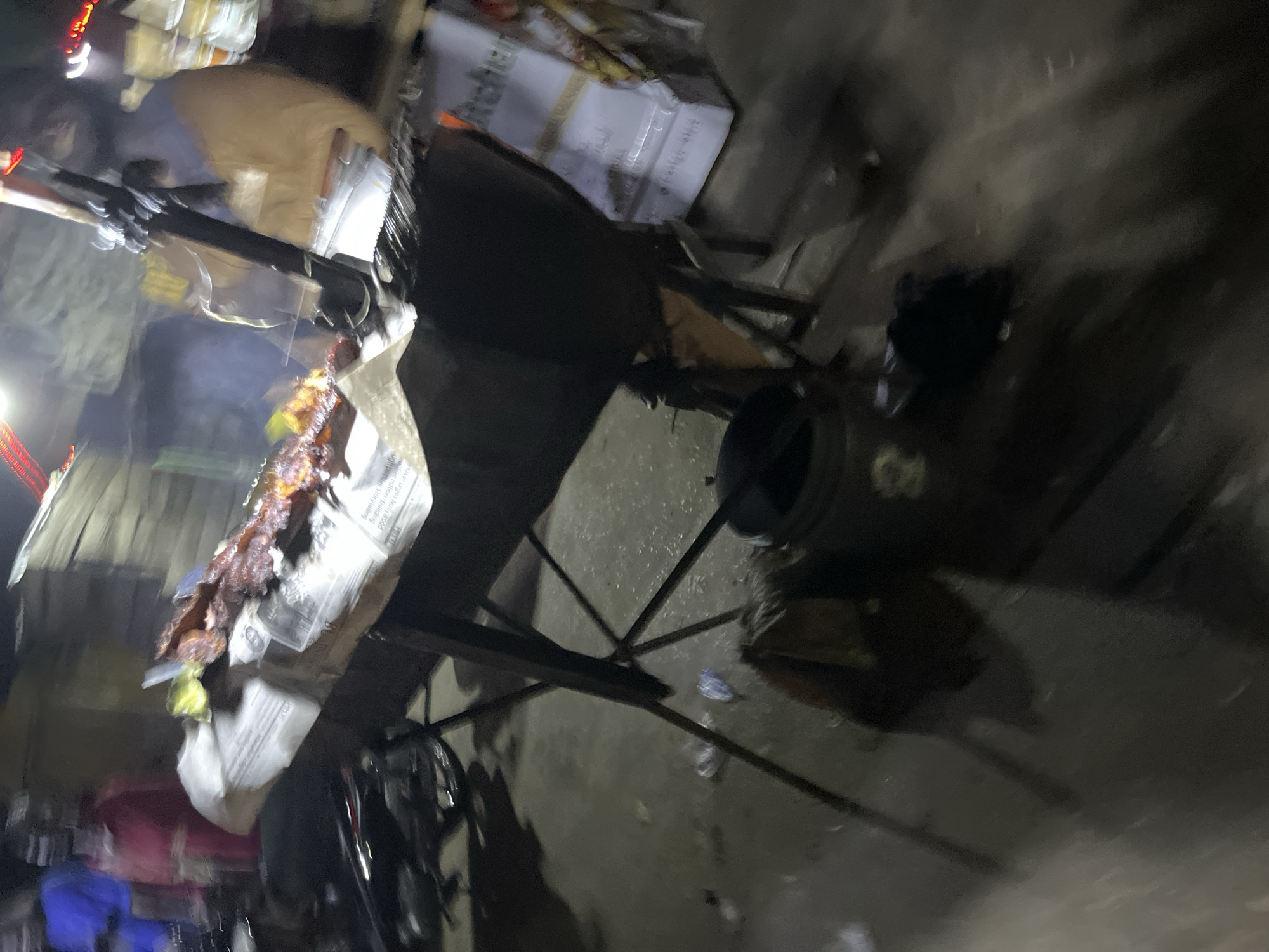A blurry, outdoor night scene shows a street food stall. A grill with food on it is visible on a table covered with newspapers. A person in a light brown garment stands behind the grill. In the background, two people in blue and red clothing are partially visible. A motorcycle is parked to the left of the stall. The ground is wet and appears to be a street. A white box with text is on the right side of the image.