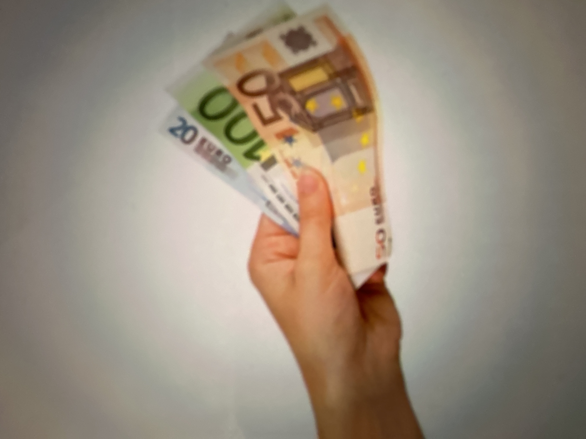 A person's hand is holding several euro banknotes: a 50 euro note, a 100 euro note, and a 20 euro note. The background is a plain, light-colored wall. The image is taken in Gelendzhik, Russia.