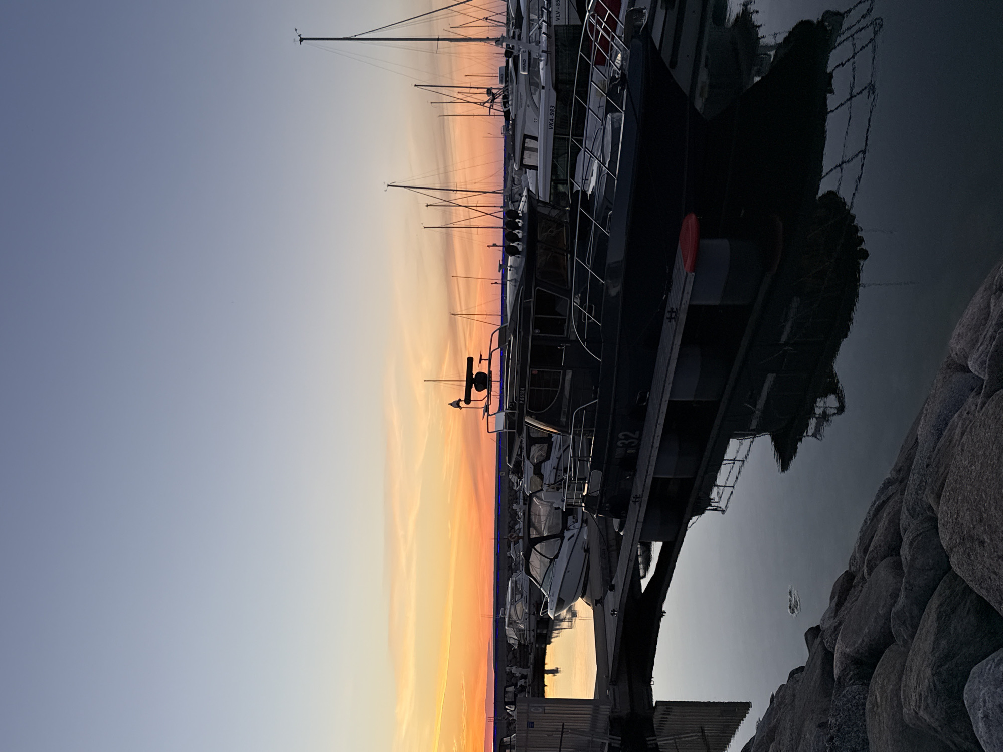 The image depicts a marina at sunset, with a clear sky transitioning from deep blue to orange and yellow hues near the horizon. Several boats are docked, with a prominent dark-colored motorboat in the foreground, displaying the number "32" on its side. Other boats, including sailboats and smaller motorboats, are visible in the background. The water reflects the boats and the colorful sky. The marina is bordered by a rocky shore on the left side of the frame. The overall scene suggests a calm evening in a coastal or waterfront area. The location is not specified in the image or the provided crops.