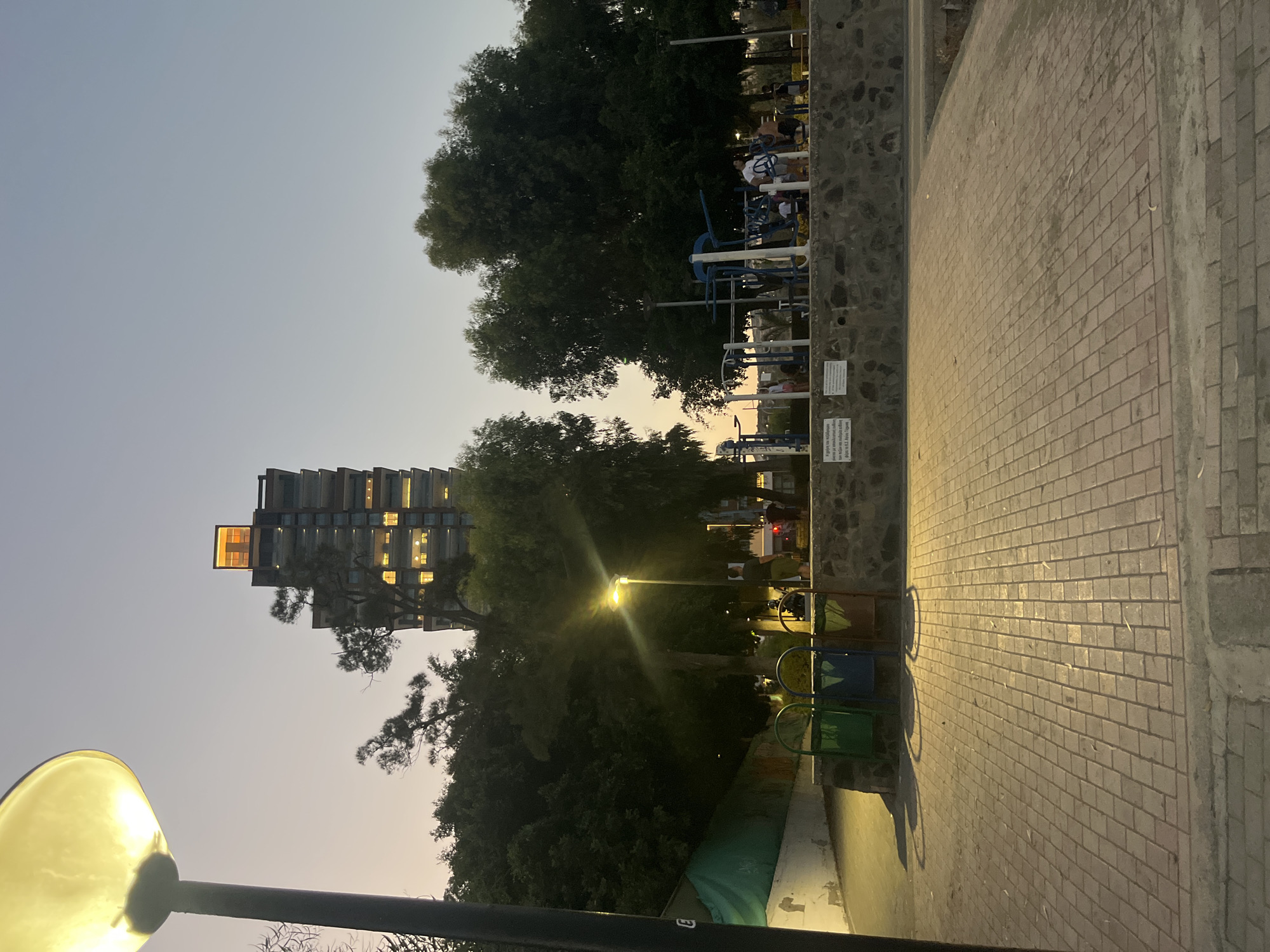 This is a low-angle, ground-level, natural light photograph taken at dusk in Agios Tychona Community, Cyprus. The scene is a paved walkway adjacent to a park area with exercise equipment and trees. In the background, a tall, modern building with illuminated windows dominates the skyline, topped with a bright orange structure. A lamppost with a lit yellow globe is visible on the left side of the frame. Three colorful trash receptacles (green, blue, and brown) are situated against a stone wall. Several people are present in the park, some appearing to use the exercise equipment. The sky is a gradient of soft blues and oranges, indicative of twilight. The ground in the foreground is paved with rectangular bricks, creating a patterned surface.
