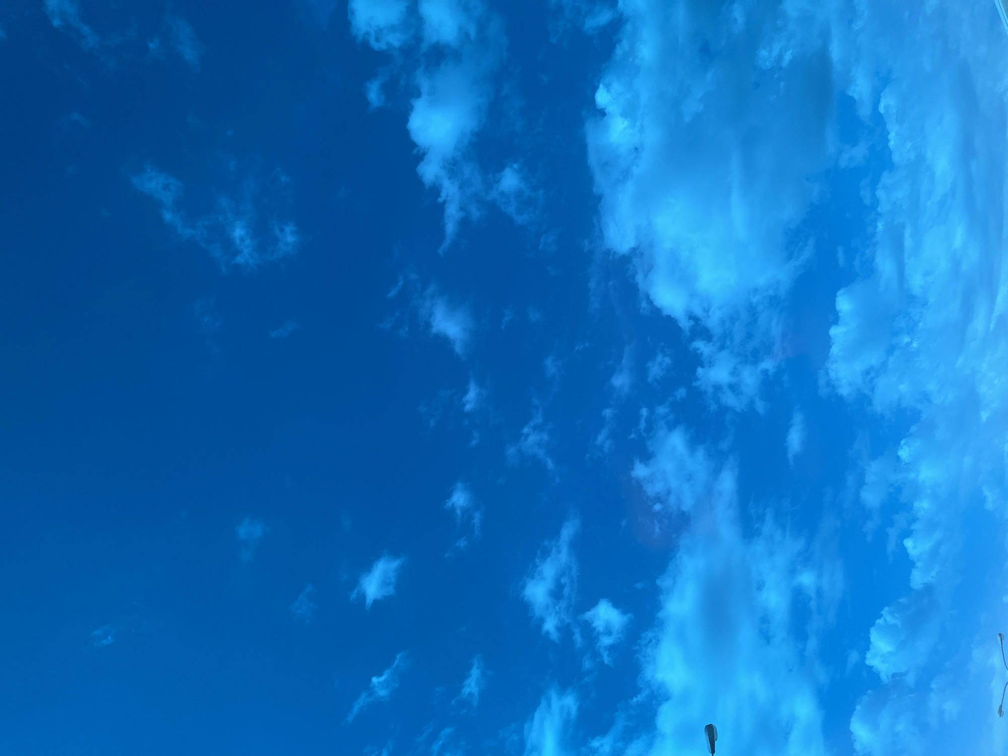 The image shows a bright blue sky with scattered clouds. The clouds range in density and shape, with some appearing as thin, wispy streaks and others as larger, more defined formations. The sky is clearest in the lower left portion of the frame. A lamppost is visible near the bottom of the frame. The photograph was taken in Tlustenkhabl, Russia.