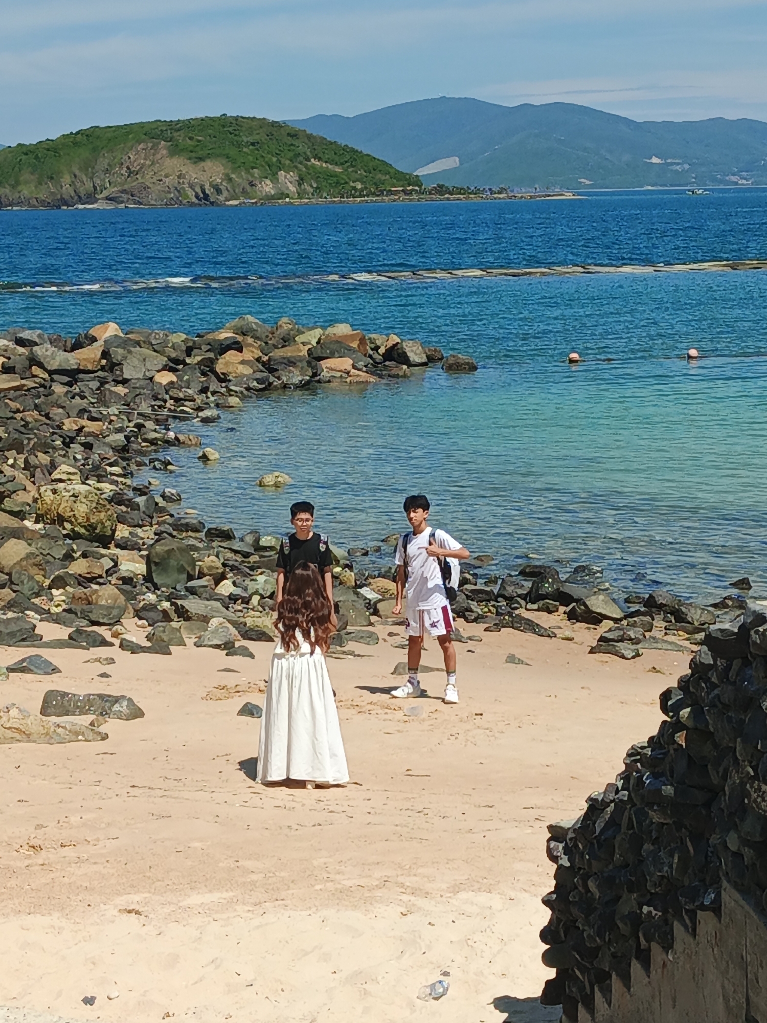 A group of three people are standing on a sandy beach next to a rocky shoreline. In the foreground, a person wearing a long, flowing white dress stands with their back to the camera. Behind them, two individuals, both appearing to be male, are standing. One is wearing a black t-shirt and glasses, while the other is wearing a white t-shirt, white shorts with red trim, and a backpack. The ocean is visible behind them, with a distant island covered in green vegetation and mountains in the background. The water is clear and blue, with some buoys floating on the surface. The scene is illuminated by bright daylight.