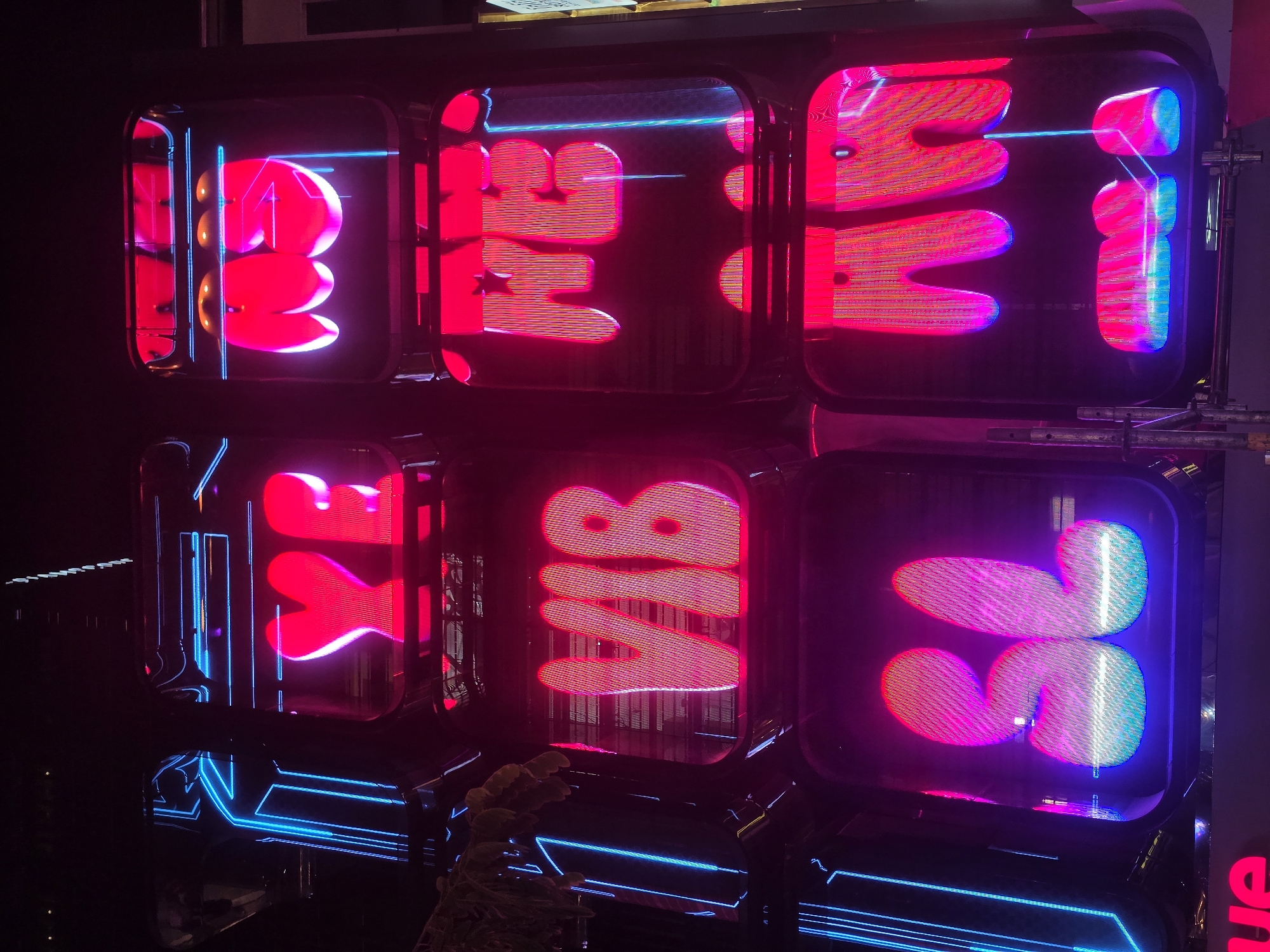 This is a nighttime photo of a neon light display in Bangkok, Thailand. The display is composed of six rectangular boxes arranged in a 2x3 grid. Each box contains a stylized neon letter or symbol in bright red and blue. From left to right, top to bottom, the boxes spell out "BET VR 36". The lighting is intense and colorful. The boxes themselves appear to be made of dark-colored, possibly metal, material. The reflections of the neon lights can be seen on the exterior surfaces. There are some reflections from nearby buildings and windows above the display. The surrounding environment is dark, with only faint glimmers of other lights and objects visible in the periphery.