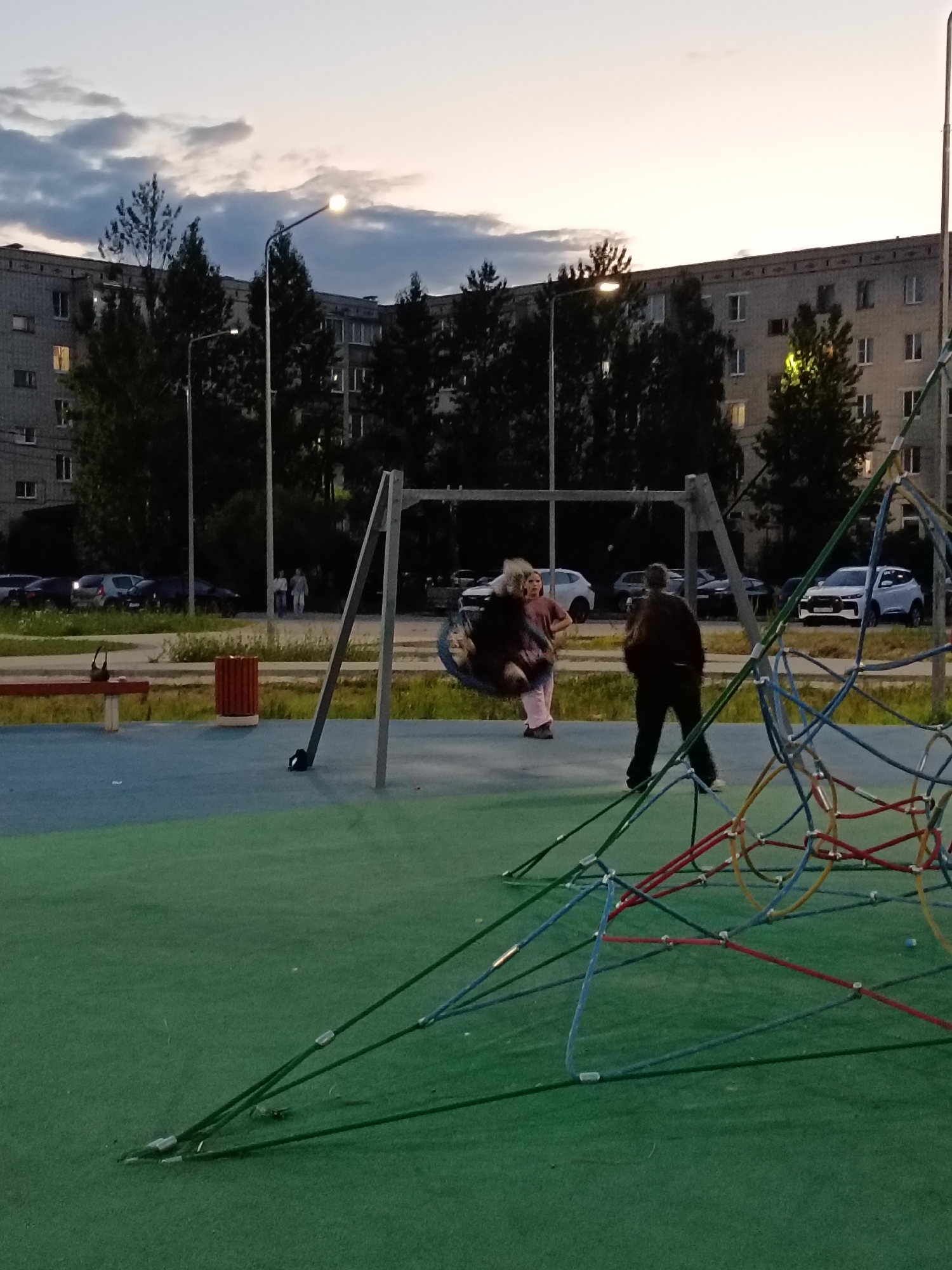 This is a vertical, full-shot image of a playground in Sudogda, Russia, captured at dusk. In the foreground, a colorful, netted climbing structure dominates the right side of the frame. The playground surface is a mix of green and blue artificial turf. In the middle ground, a swing set is visible with two people on it: a woman in a pink outfit is on the swing, and a man in a dark jacket stands nearby. Further back, a multi-story apartment building with numerous windows is seen behind a line of trees. Several cars are parked in front of the building, and streetlights illuminate the scene. The sky is overcast with soft clouds, suggesting the end of daylight.