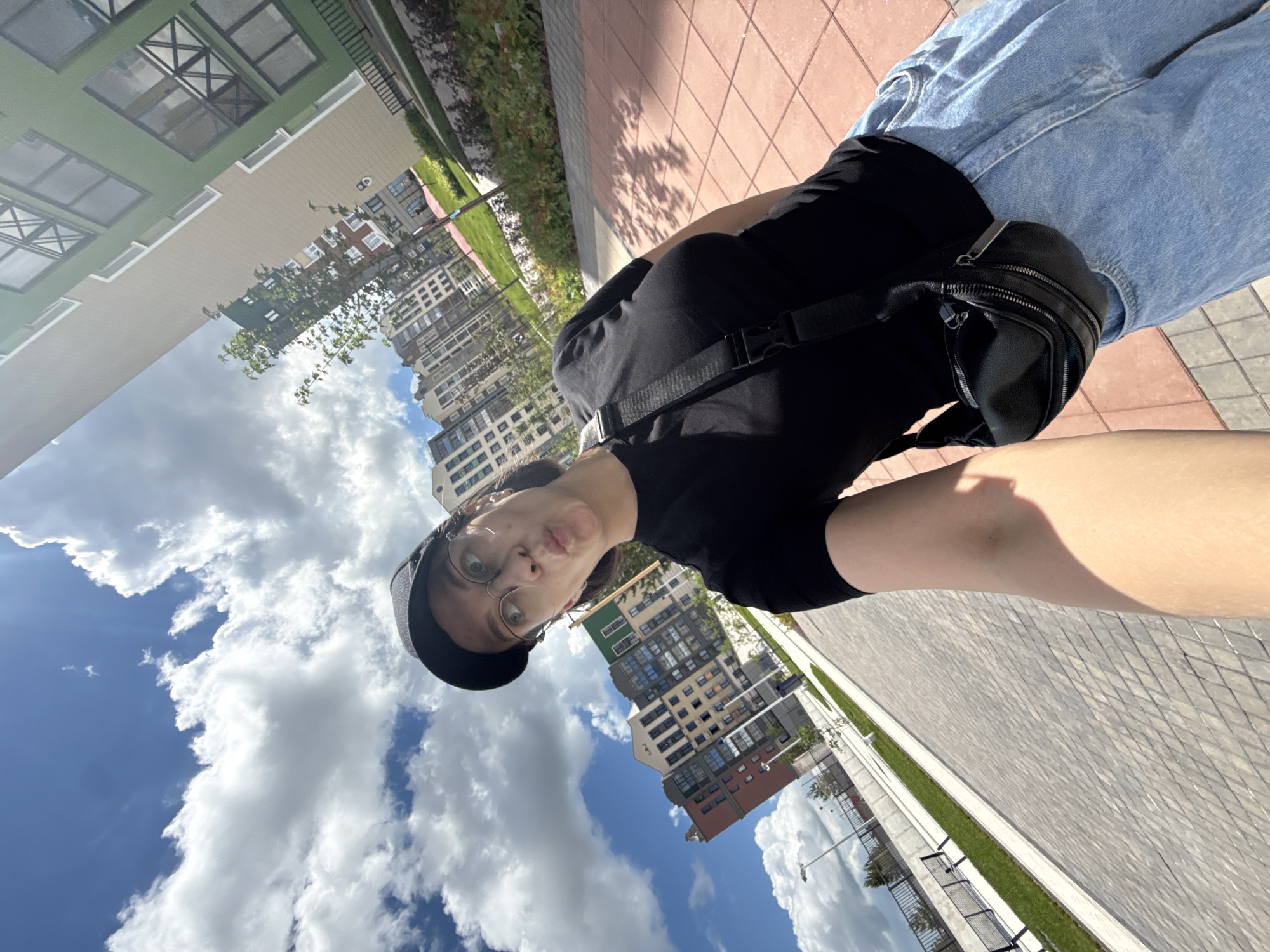 A young woman with glasses and a black baseball cap takes a selfie while walking outdoors. She is wearing a black t-shirt, light blue jeans, and a black fanny pack across her chest. She is making a kissing face at the camera. In the background, there are modern apartment buildings with colorful facades and a blue sky with fluffy clouds. The ground is paved with interlocking bricks. The scene appears to be in a newly developed residential area in Izhevsk, Russia, possibly a courtyard or a street within a housing complex.