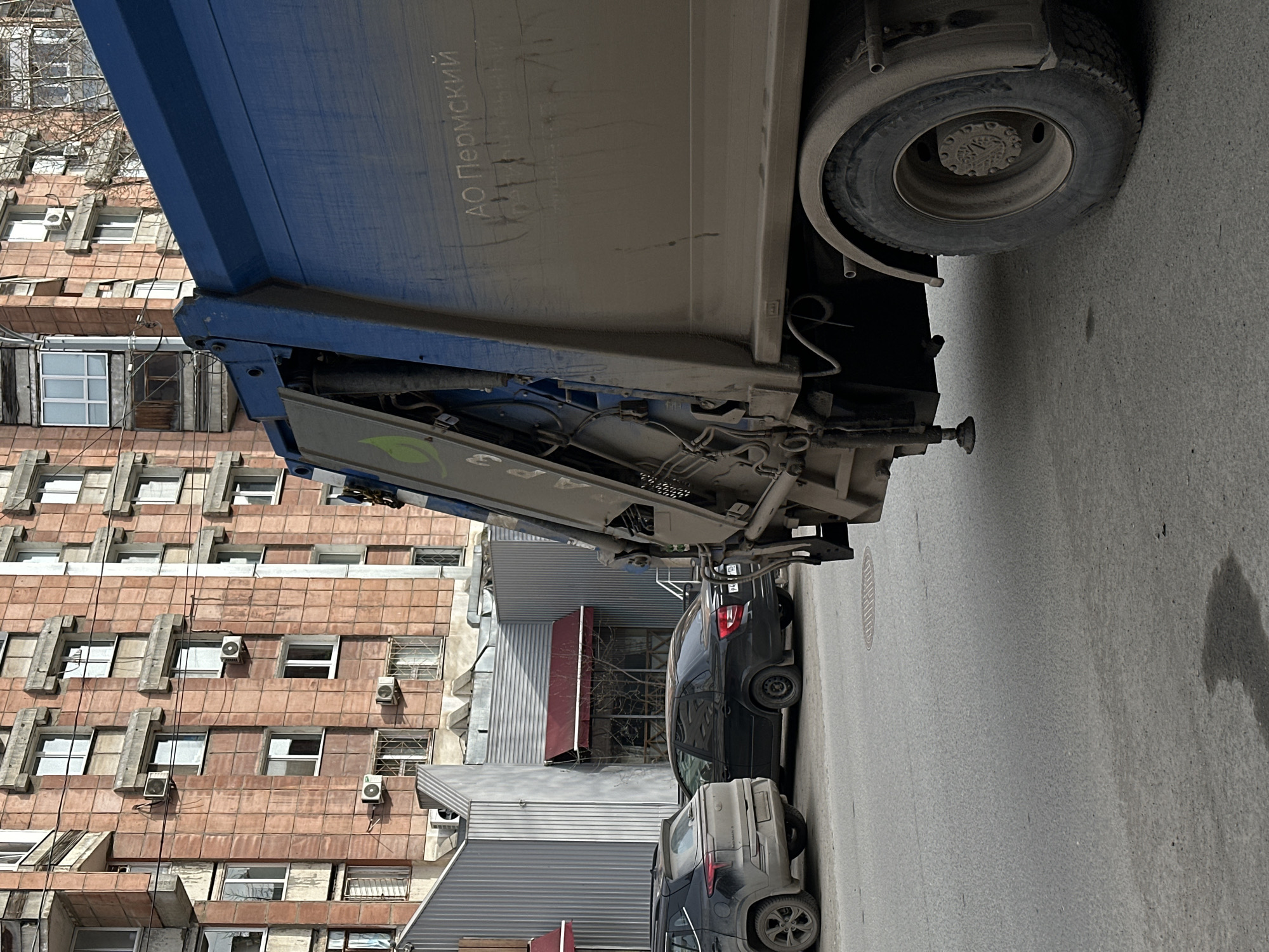 The image shows a parked garbage truck in an urban setting, possibly in Perm, Russia, as indicated by the Cyrillic text "АО Пермский" visible on the side of the truck. The truck, predominantly blue and beige, is parked parallel to the curb on an asphalt street. Several parked cars, including a dark sedan and a grey SUV, are aligned along the street. A multi-story building with a facade of red-brown tiles and numerous windows with air conditioning units is visible in the background. Additionally, the upper portion of a grey metal building with a red awning is situated nearby. The street appears to be in a well-populated area.