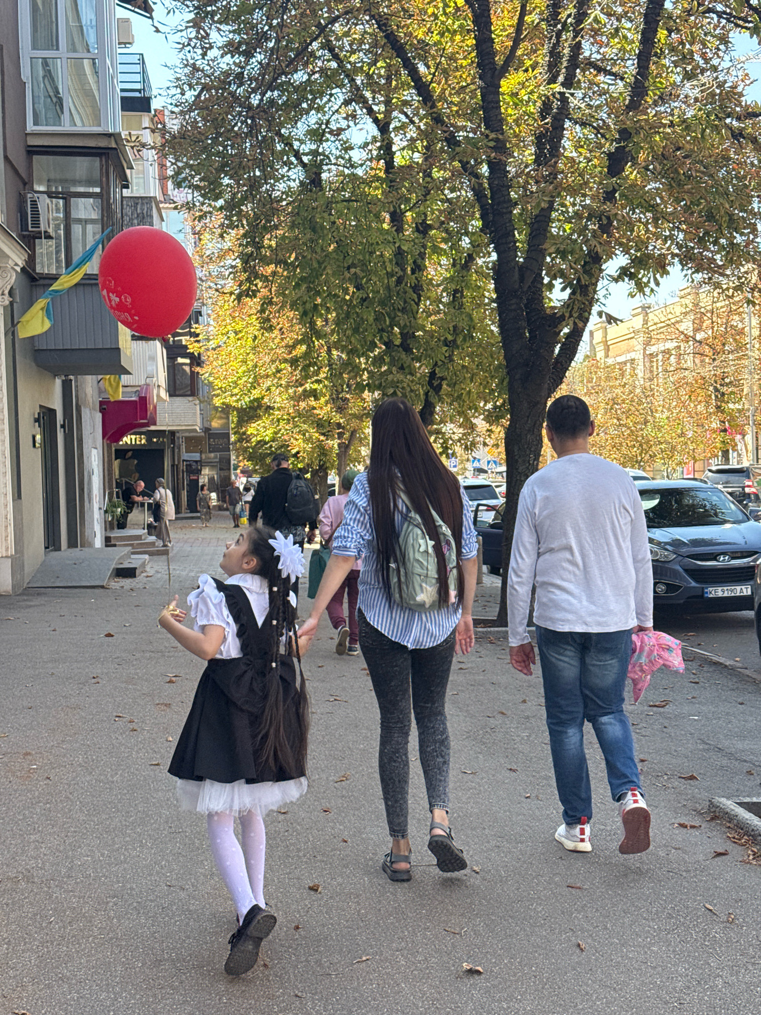 The image depicts a street scene in Kryvyi Rih, Ukraine, on a sunny day with a clear blue sky. In the foreground, a young girl in a black and white school uniform with white bows in her hair looks up at a large red balloon, which is tethered to a building on the left. She is holding a small object in her right hand. Behind her, a woman with long dark hair, wearing a striped shirt and a backpack, walks away from the camera, holding the girl's left hand. To the right of the woman, a man in a white long-sleeved shirt and blue jeans also walks away from the camera, carrying a pink bundle in his right hand.
The street is paved with asphalt and lined with a sidewalk. Buildings with multiple floors are visible on both sides of the street, some with balconies and windows. Trees with yellowing leaves, suggesting autumn, line the sidewalk, casting shadows on the ground. Several cars are parked or driving on the street. A Ukrainian flag is visible on the side of a building on the left. The overall atmosphere appears to be casual and pedestrian.