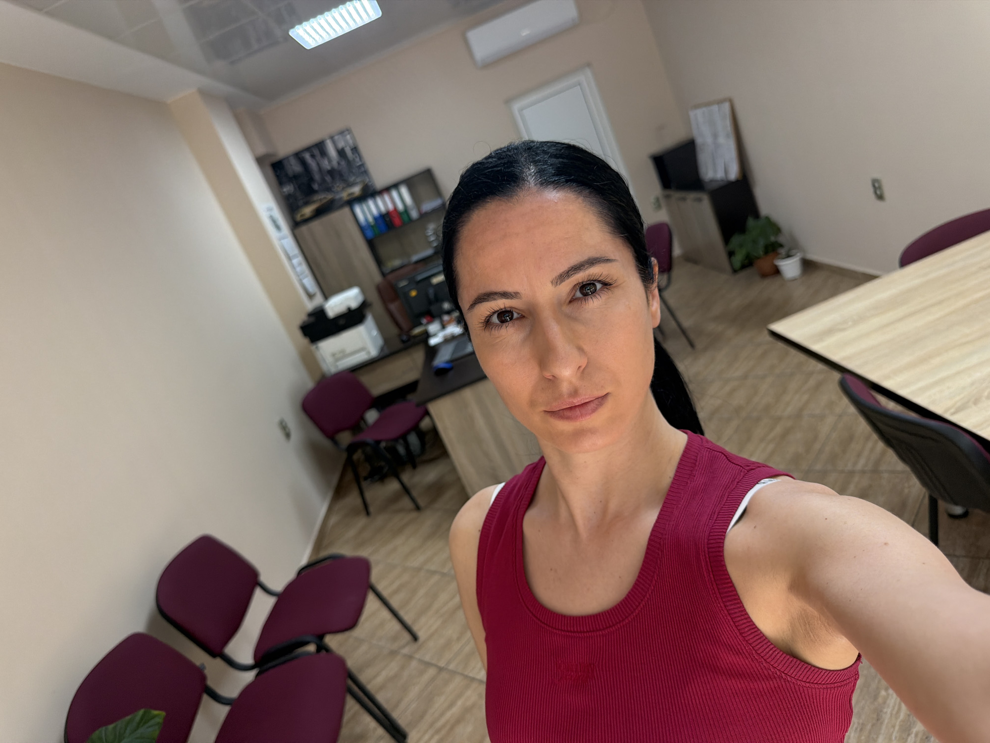 A woman with dark hair pulled back captures a selfie in what appears to be an office or meeting room in Pleven, Bulgaria. She is positioned in the foreground, facing the camera with a neutral expression. She wears a ribbed maroon tank top, with a white strap visible on her left shoulder.
The background reveals a well-lit indoor environment with light beige walls and a light-colored tiled floor. To the left and behind the woman, an office area is visible, featuring a dark desk, a filing cabinet with colorful binders, and a printer. Several burgundy office chairs with black frames are arranged around this desk and further into the room.
To the right, a large wooden table with a black edge, also accompanied by burgundy chairs, suggests a meeting or collaboration space. Above, fluorescent strip lights illuminate the room, and an air conditioner is mounted on the wall. A framed image or poster is visible on the wall behind the desk. A couple of potted plants are also discernible on the floor in different parts of the room, adding a touch of greenery. The overall setting is tidy and functional, indicative of a professional workspace during working hours. No explicit text is clearly visible in the image.
