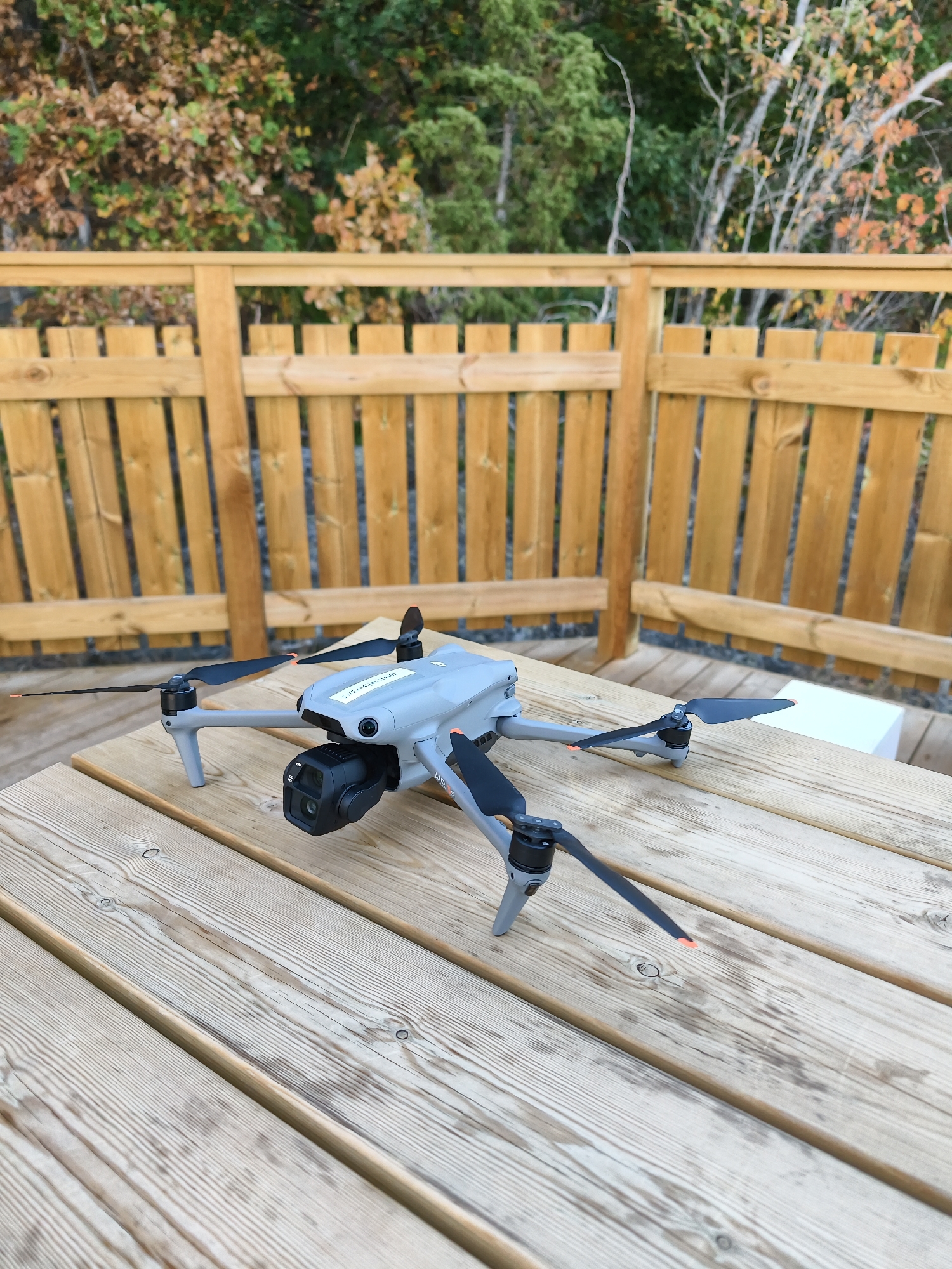 A gray DJI Mavic 3 drone is positioned on a light-colored wooden deck. The drone has four black propellers, two of which are extended forward and two are folded back. The camera gimbal with a black lens is visible on the front of the drone. A white sticker with text is attached to the top of the drone. Behind the drone, a light brown wooden fence with vertical planks is visible. Beyond the fence, there are trees with green and orange foliage, suggesting it might be autumn. The drone is the primary subject, with the wooden deck and fence serving as the immediate environment. The scene appears to be outdoors, possibly in a park or a residential area with a natural setting, which could be consistent with Söderköping, Sweden, a town known for its natural beauty and proximity to forested areas and waterways. There are no people or active interactions depicted in the image.