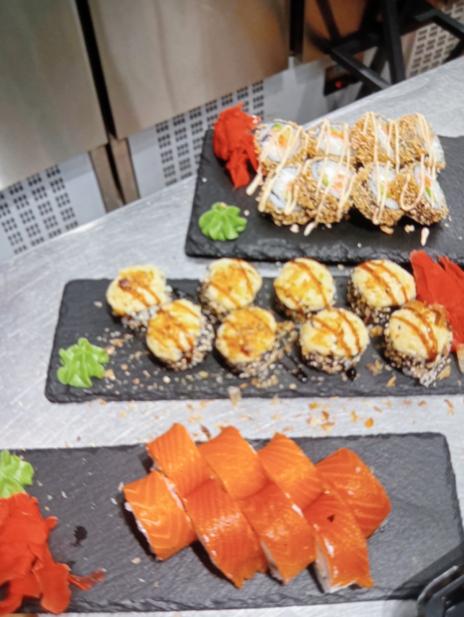The image displays an assortment of sushi rolls arranged on dark slate platters. In the top platter, there are several sushi rolls that appear to be deep-fried, coated in a crispy exterior, and drizzled with a creamy sauce. Red pickled ginger and a dollop of green wasabi are placed to the left of these rolls. Below, a second platter holds more sushi rolls, these with a golden-brown, seared topping, also drizzled with sauce, and accompanied by wasabi. The bottom platter features slices of raw salmon arranged in a fan shape, with red pickled ginger and wasabi to the left. The scene is set on a metallic surface, possibly a kitchen counter or a restaurant preparation area, with a blurred background that suggests an indoor setting, possibly a commercial kitchen with stainless steel appliances. The lighting is artificial and somewhat dim, casting a slight sheen on the food. The overall impression is of a carefully prepared and presented sushi meal. There is no visible text in the image.