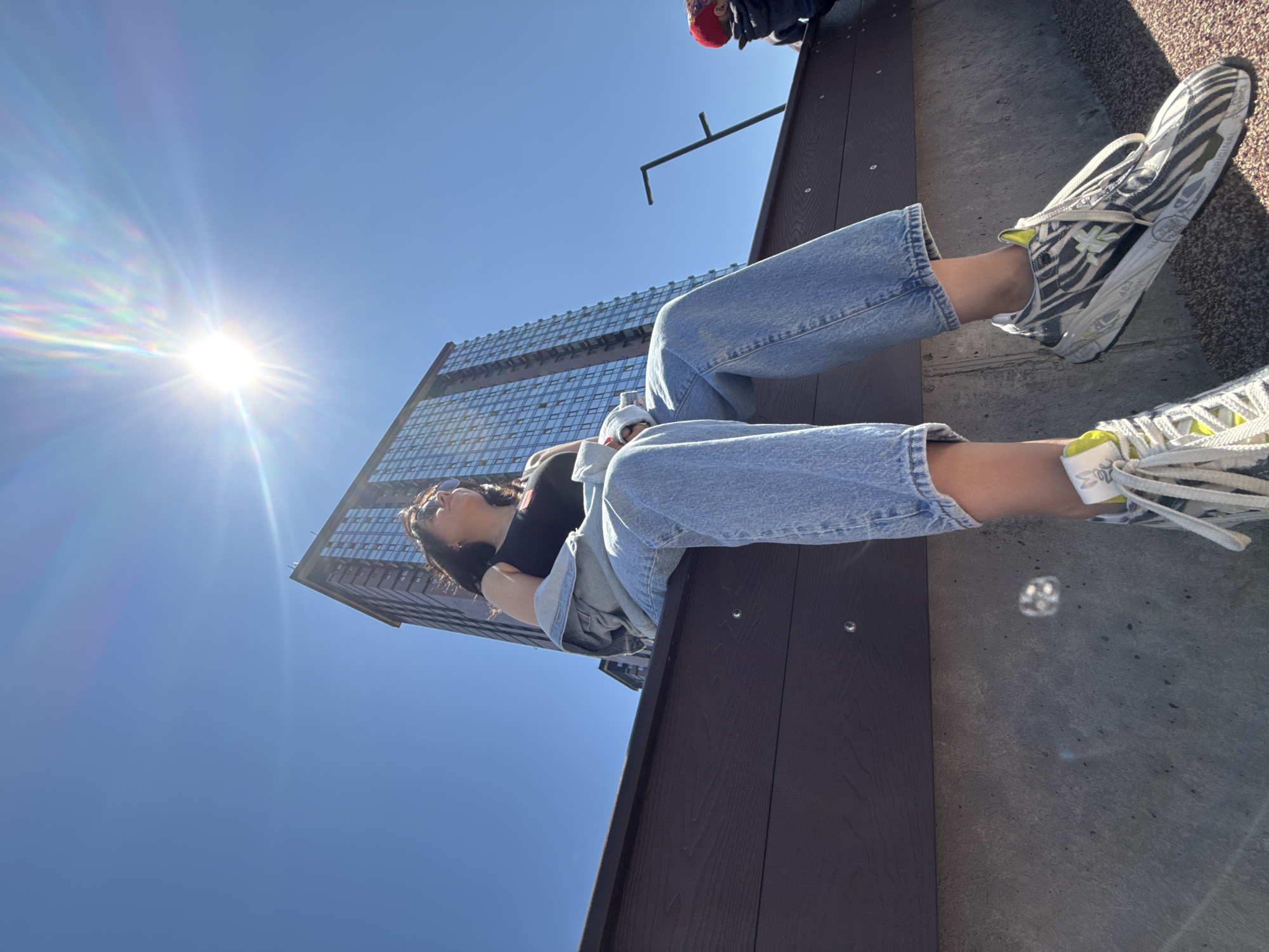 A person is reclining on a park bench in what appears to be an urban setting. The person is wearing blue jeans, a black top, a light jacket, and sneakers with a zebra-stripe pattern. They are lying back with their hands behind their head, looking up towards the sky. Above them is a clear blue sky with the sun shining brightly. A tall building is visible in the background. There are visible rivets on the park bench. There is a round glare reflection on the ground behind the bench. The location of the photo is Vladivostok, Russia.